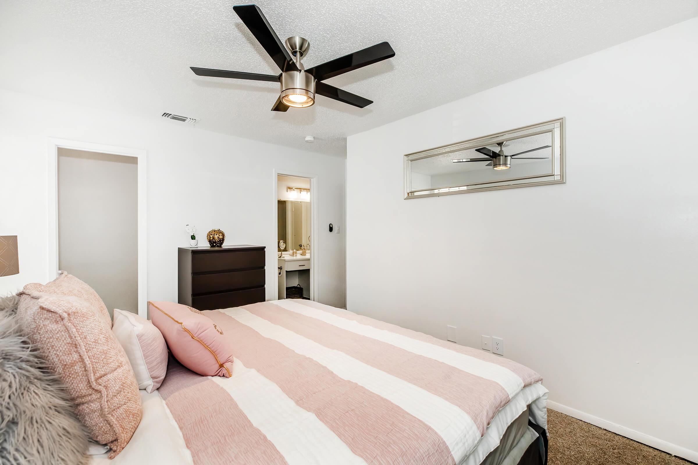 A modern bedroom featuring a neatly made bed with pink and white striped bedding. There's a plush throw pillow, a dresser, and a mirror on the wall reflecting a ceiling fan. A door leads to a bathroom area, and the carpeted floor adds warmth to the space. White walls create a bright and airy atmosphere.