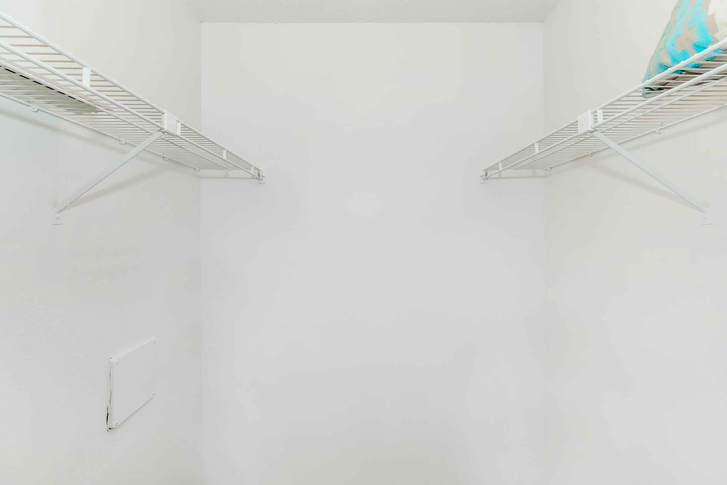 A clean, empty closet with two wire shelves on either side. The walls and ceiling are painted white, creating a bright and open feel. There is a small access panel on the left wall, and a light blue item is placed on one of the shelves.