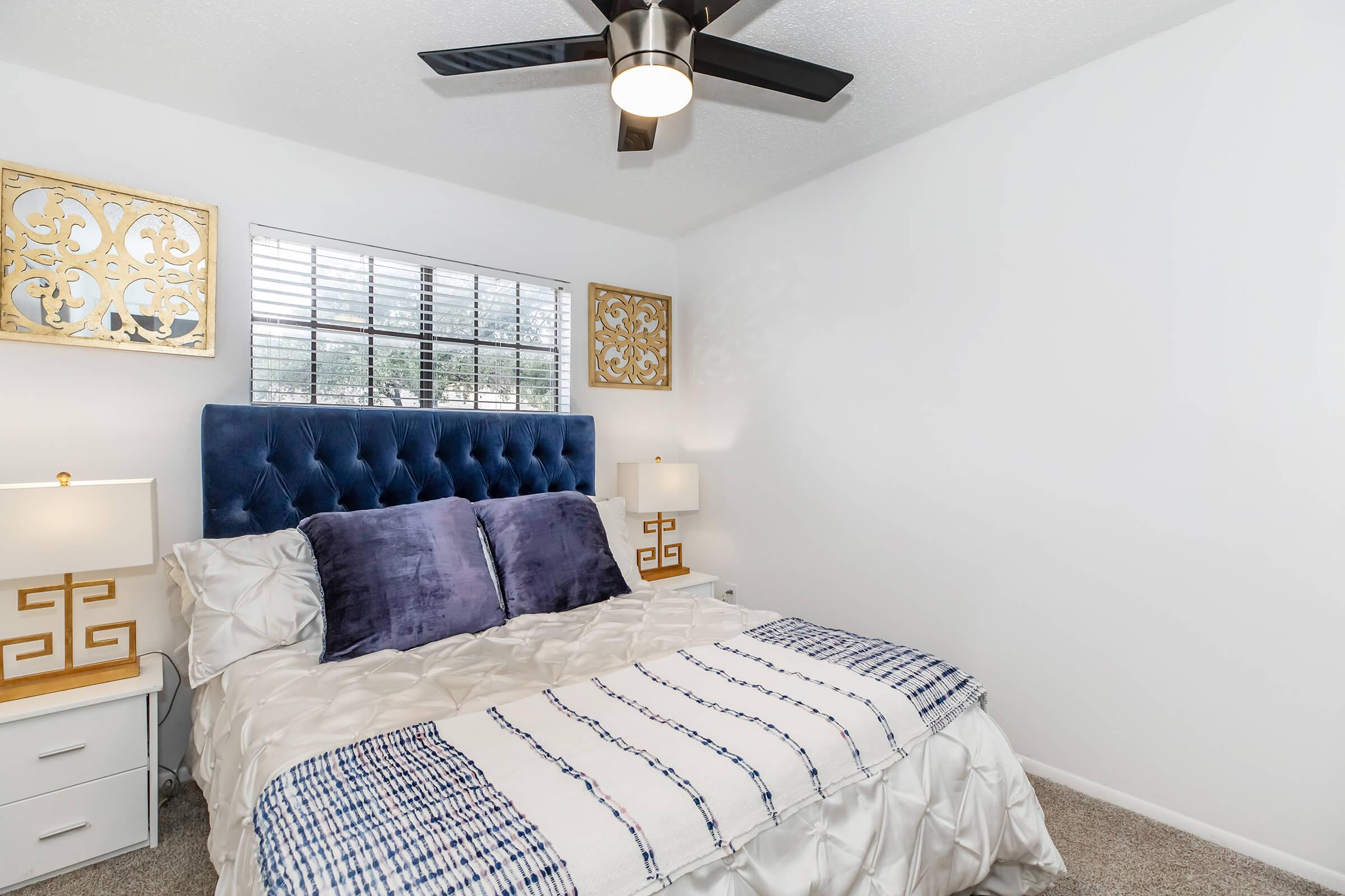 A cozy bedroom featuring a blue tufted headboard with two plush pillows, a neatly made bed with a white comforter and a blue and white patterned throw. Two decorative lamps on nightstands provide soft lighting, and a ceiling fan is mounted above. Decorative gold wall art complements the room's modern design.