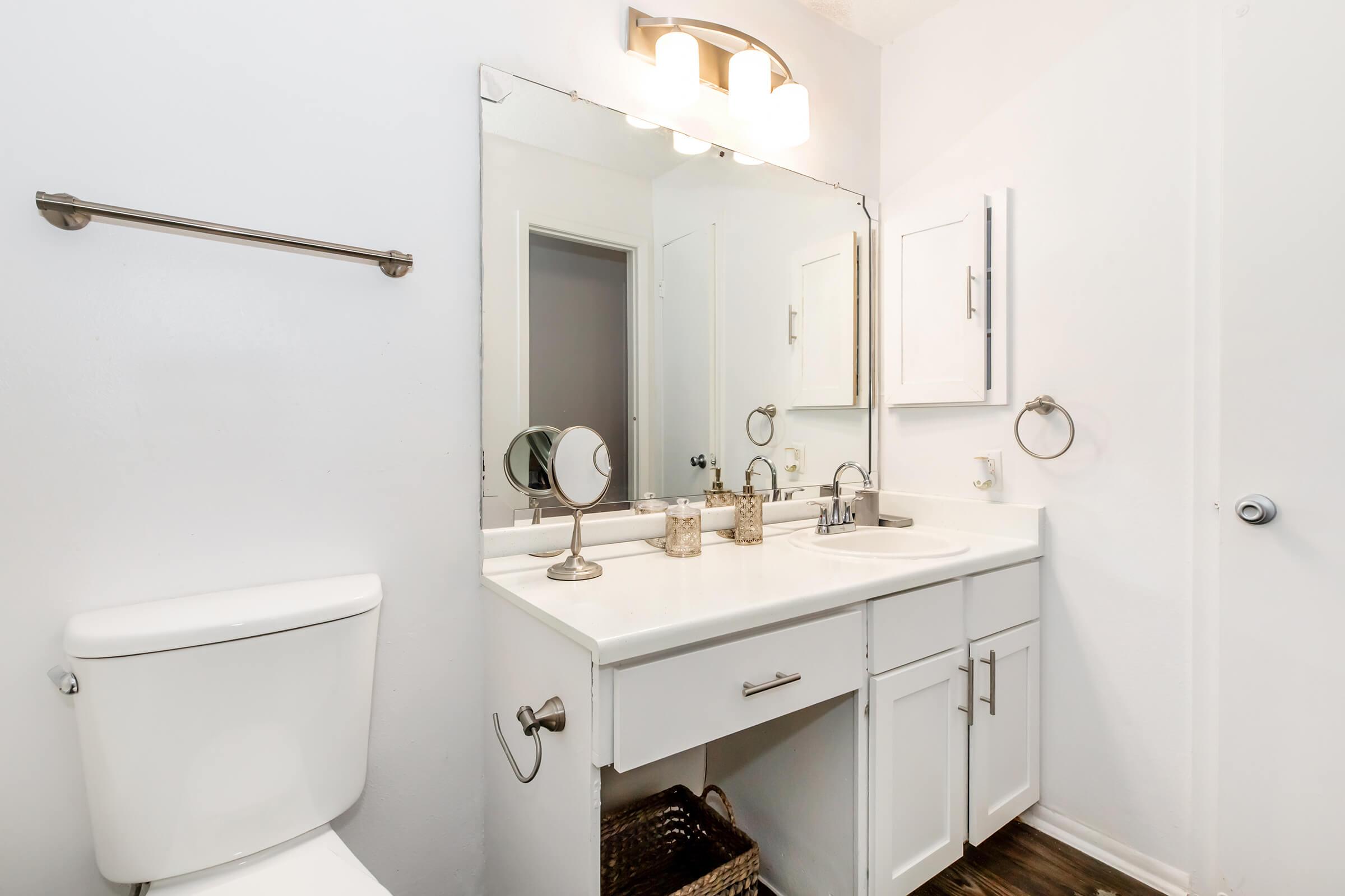 A clean and modern bathroom featuring a white vanity with a sink, an illuminated mirror, a round makeup mirror, a towel rack, and a cabinet. The toilet is situated to the left, with neutral-colored walls and wooden flooring creating a bright and inviting atmosphere.