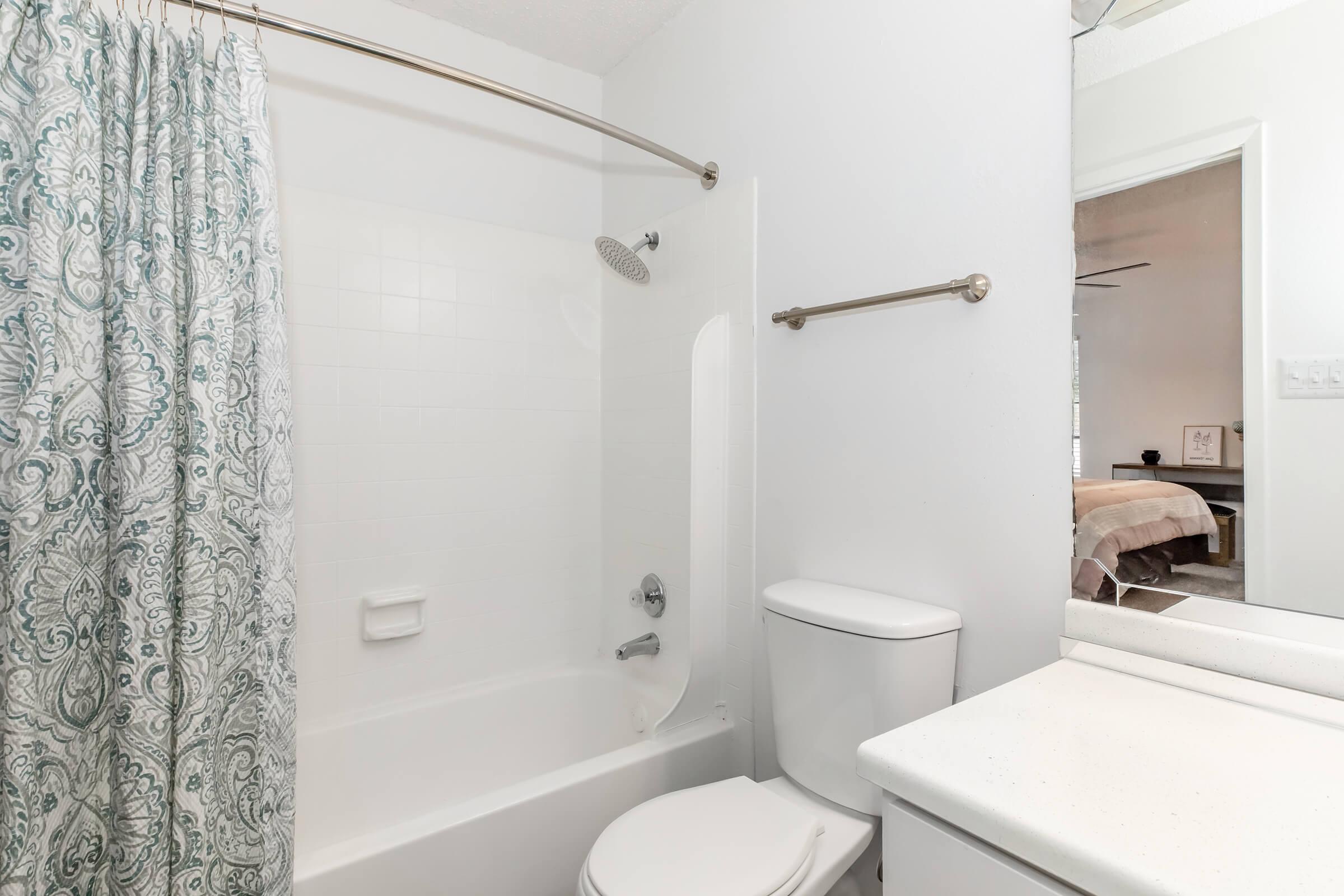 A clean, modern bathroom featuring a bathtub with a shower curtain, a toilet, and a sink with a white countertop. The walls are painted white, and there is a towel rack. In the background, a glimpse of a bedroom can be seen through the door, showcasing a bed with neutral-colored bedding.