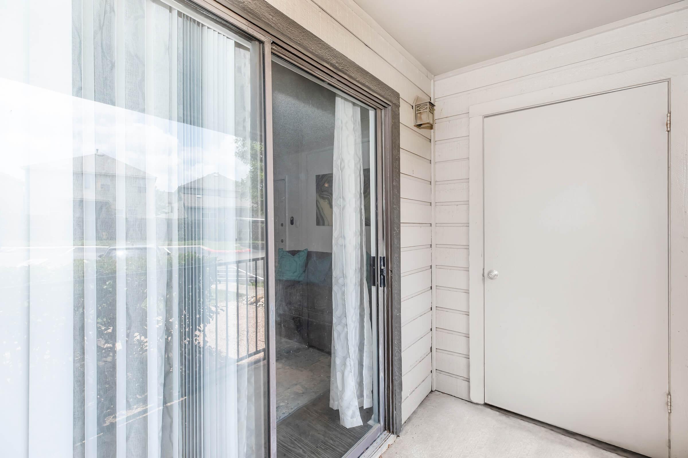 A modern entryway with sliding glass doors revealing a living space. The interior features a blue couch and decorative items. Natural light streams in through the sheer white curtains, and a solid white door is visible on the right, complementing the light-colored wood siding of the exterior.