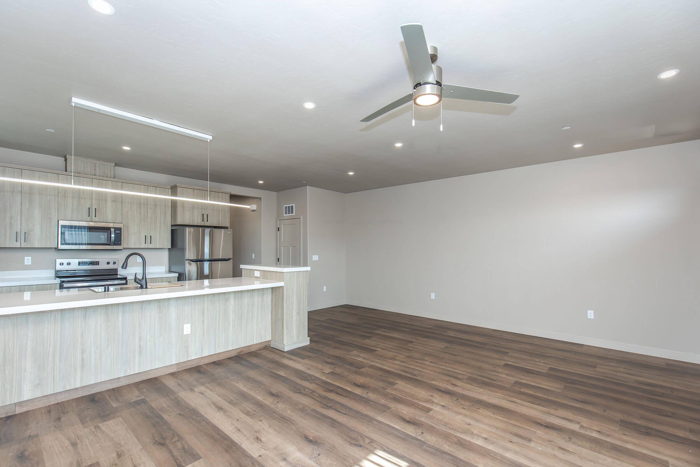 Modern apartment interior featuring an open floor plan, light-colored walls, a spacious living area with laminate wood flooring, a ceiling fan, and a kitchen bar with stainless steel appliances. Natural light illuminates the space from overhead lighting fixtures.