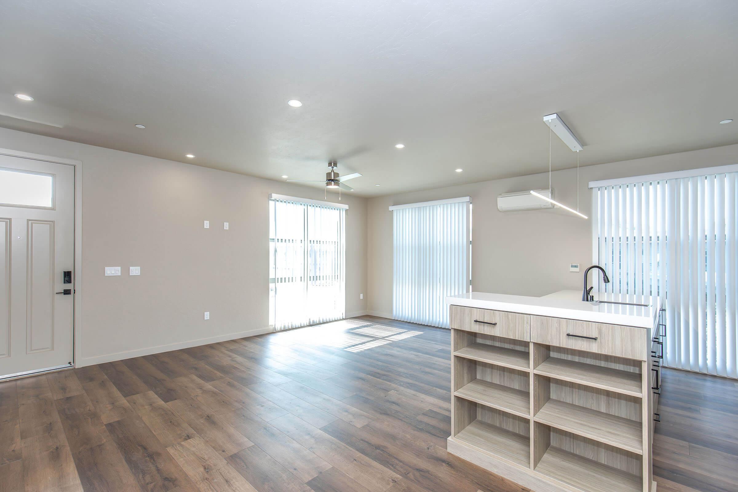 A modern, well-lit living space featuring an open floor plan. The room has large windows with vertical blinds, a ceiling fan, and wood laminate flooring. A stylish kitchen island with shelving is visible, along with a door leading outside. The overall color scheme is neutral, creating a bright and airy atmosphere.