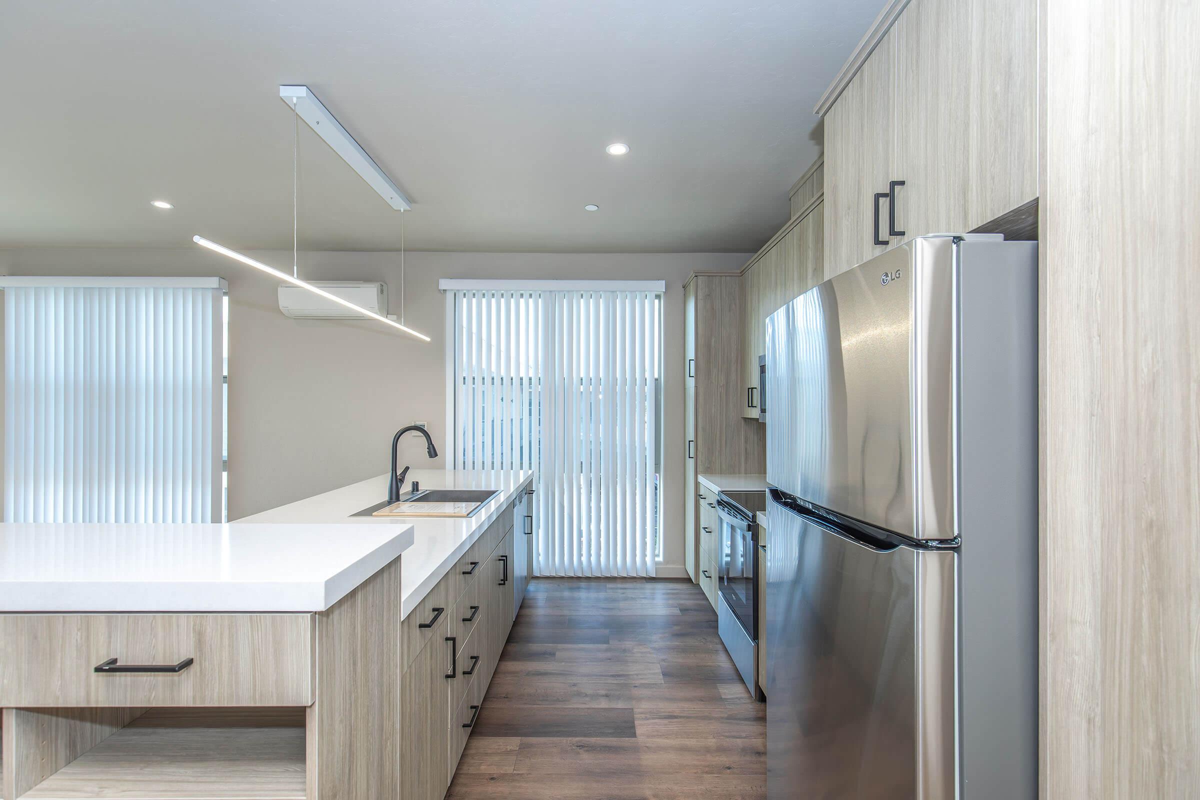 Modern kitchen featuring light wood cabinetry, a large white countertop, and stainless steel appliances. A sleek pendant light hangs above the kitchen island, and there are vertical blinds covering a sliding door that leads outside. The floor is a warm-toned wood laminate, creating an inviting atmosphere.