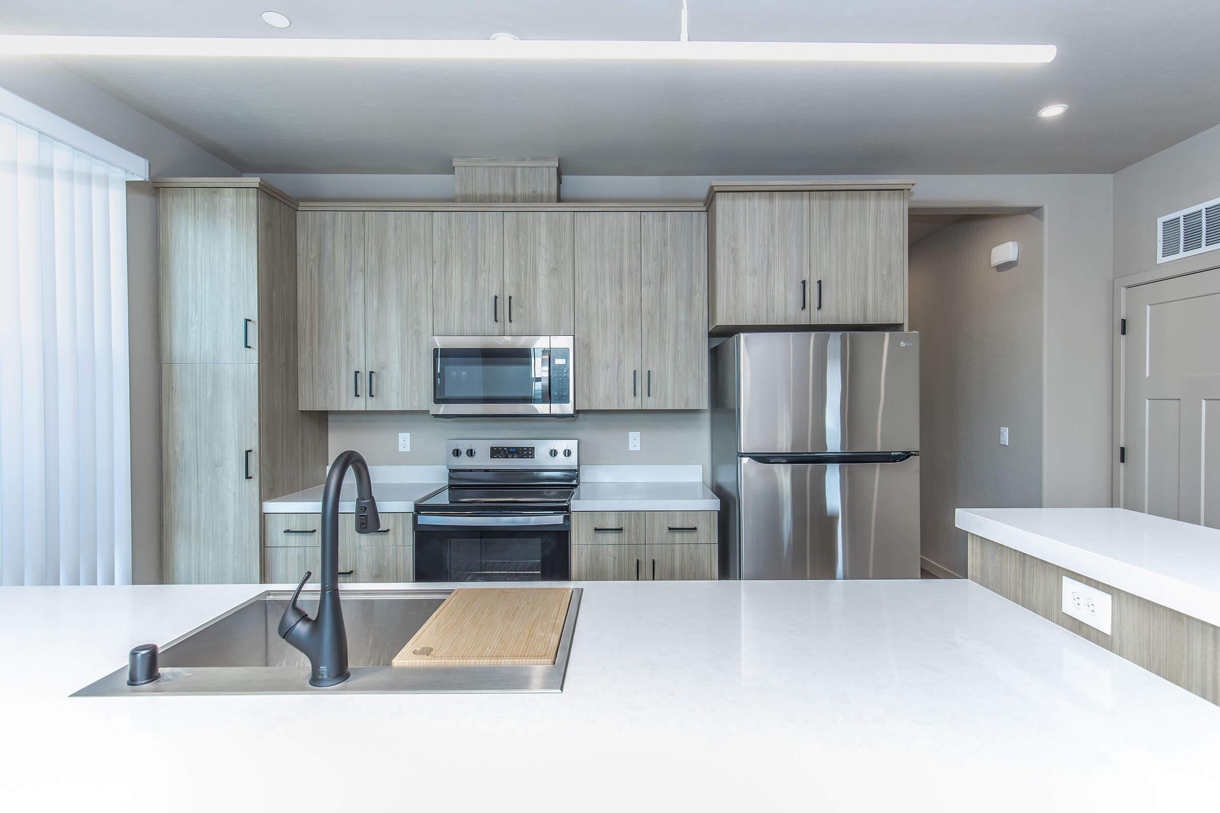 Modern kitchen featuring light wood cabinetry, stainless steel appliances including a microwave and fridge, a sleek black faucet, and a spacious white countertop. Soft lighting illuminates the area, creating a clean and inviting atmosphere.