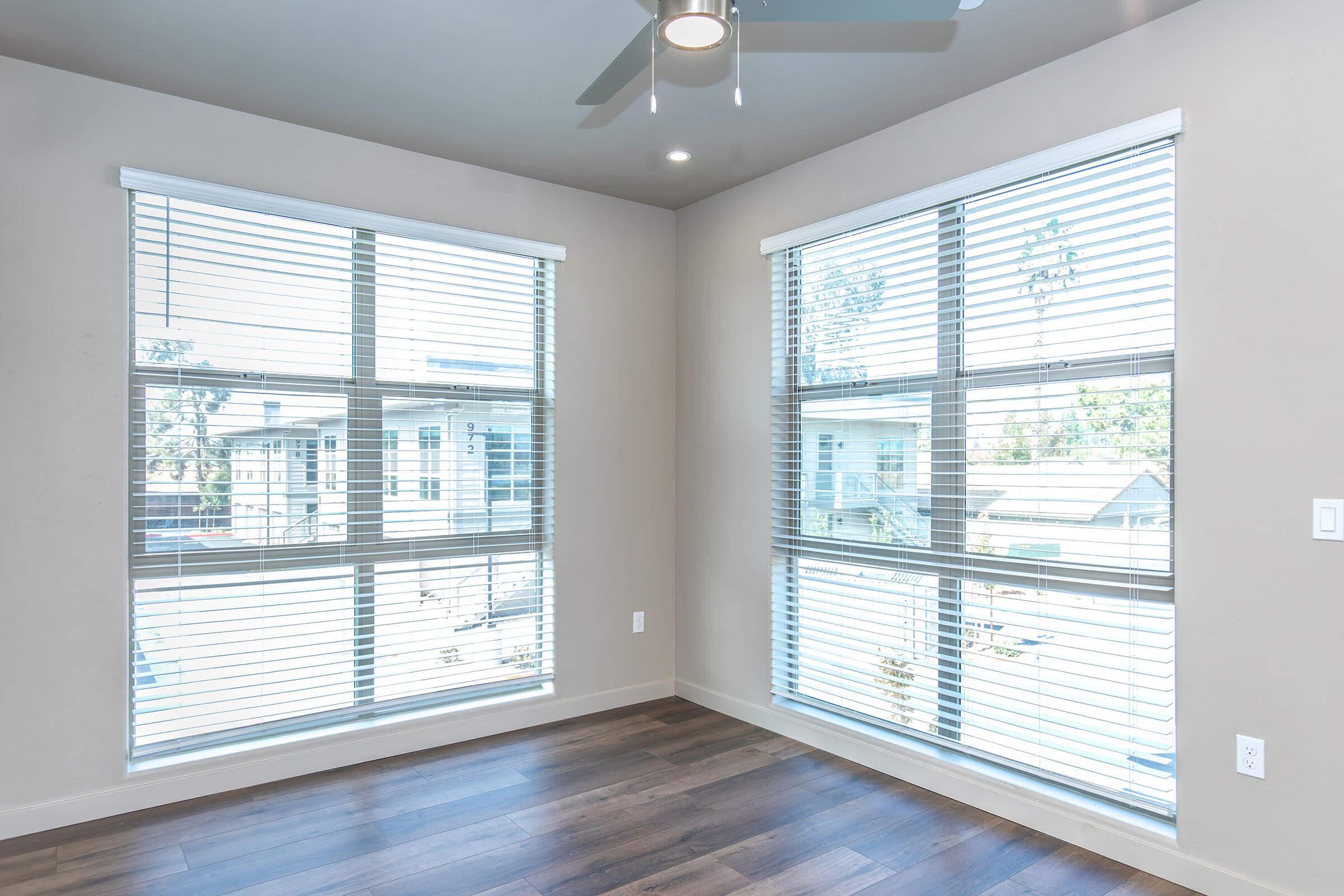 A bright room featuring two large windows with white blinds, hardwood flooring, and light-colored walls. The ceiling has a modern ceiling fan, and outside the windows, greenery and buildings are visible in the background. The space appears open and airy, ideal for natural light.