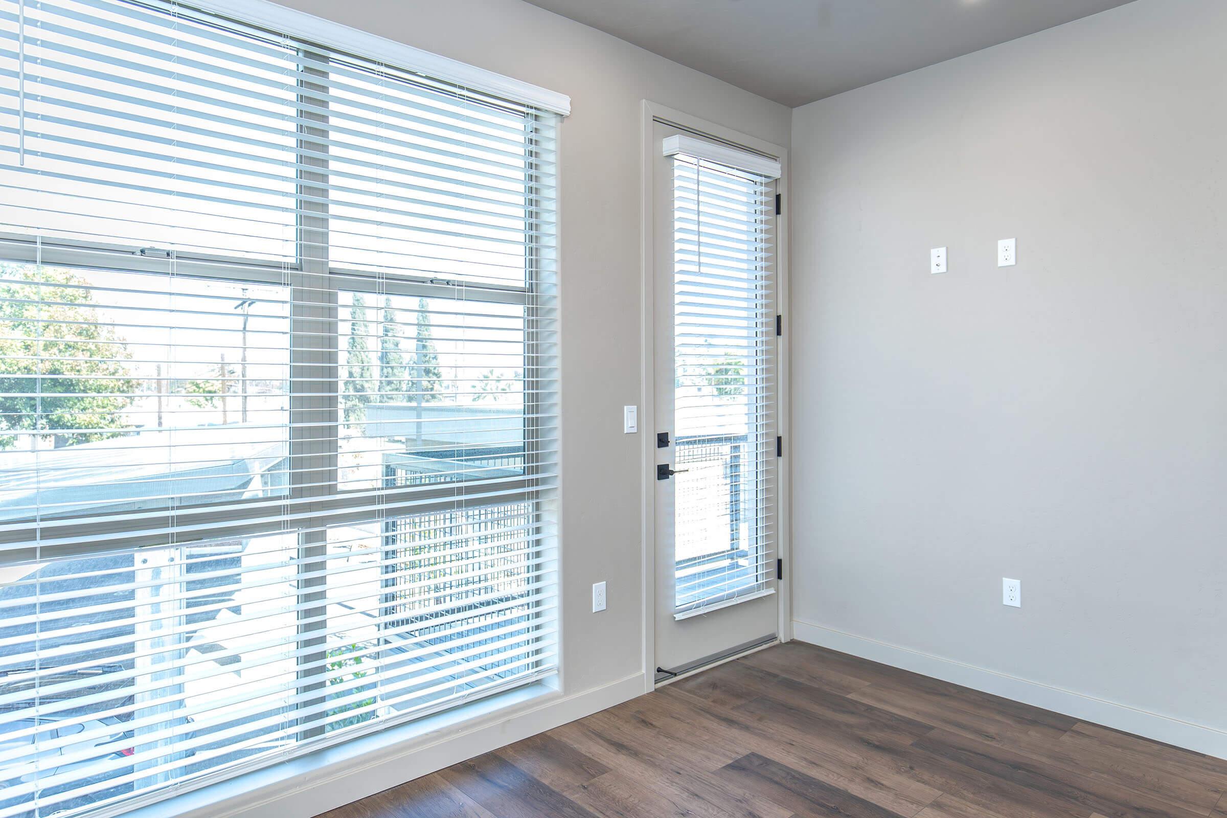 Bright, modern room with large windows featuring white blinds, allowing natural light to filter in. The walls are light-colored, and there’s a sliding glass door leading to a balcony. The floor is made of polished hardwood, enhancing the contemporary feel of the space.