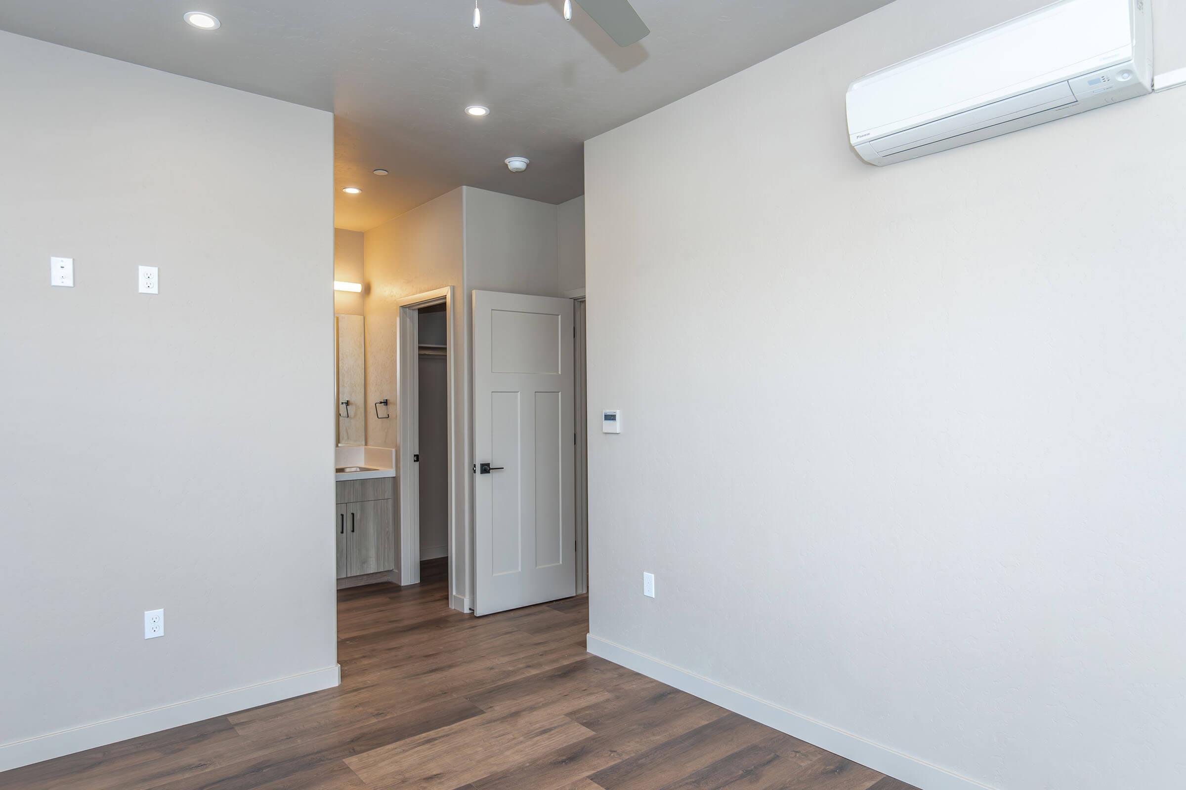 Interior of a modern room featuring light-colored walls, hardwood flooring, and recessed ceiling lights. A door leads to a bathroom area in the background, while an air conditioning unit is mounted on the wall. The space appears bright and newly renovated, with minimal furnishings.