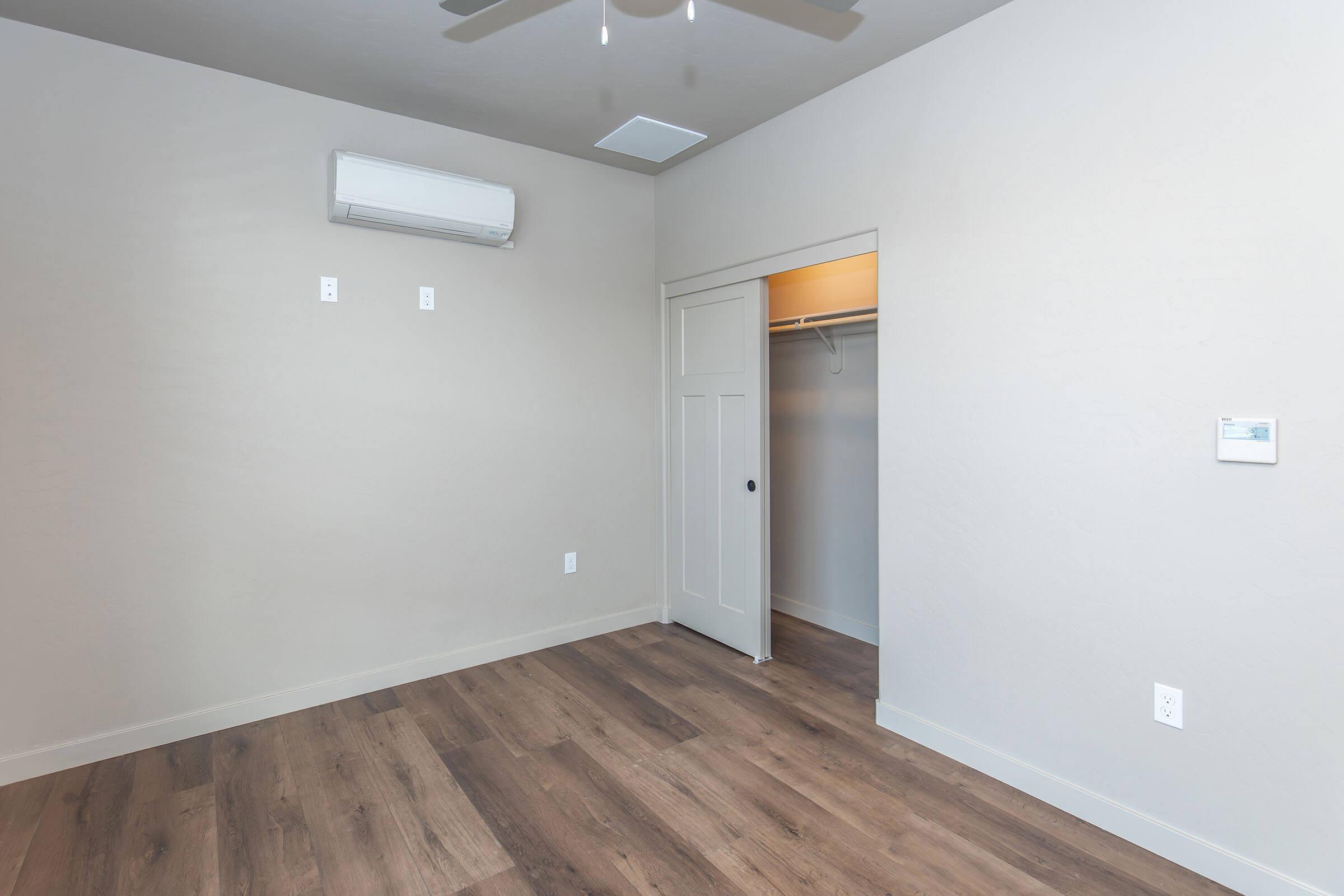 A spacious, empty room with light-colored walls and a wooden floor. An air conditioning unit is mounted on the wall, and a white closet door leads to a closet space. The room is bright and minimally decorated, featuring a ceiling fan and a thermostat on the wall.