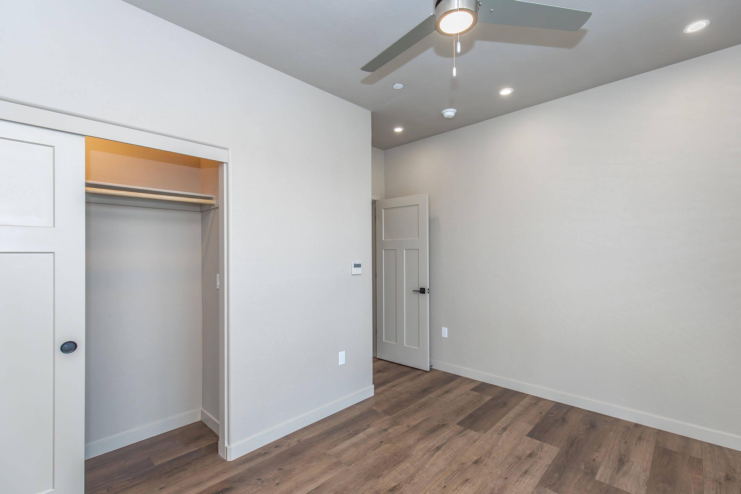 A light-colored room with a wooden floor, featuring a ceiling fan. There’s an open closet on the left and a closed door on the right. The walls are painted in a soft hue, and there are recessed lights in the ceiling, creating a bright and airy atmosphere.