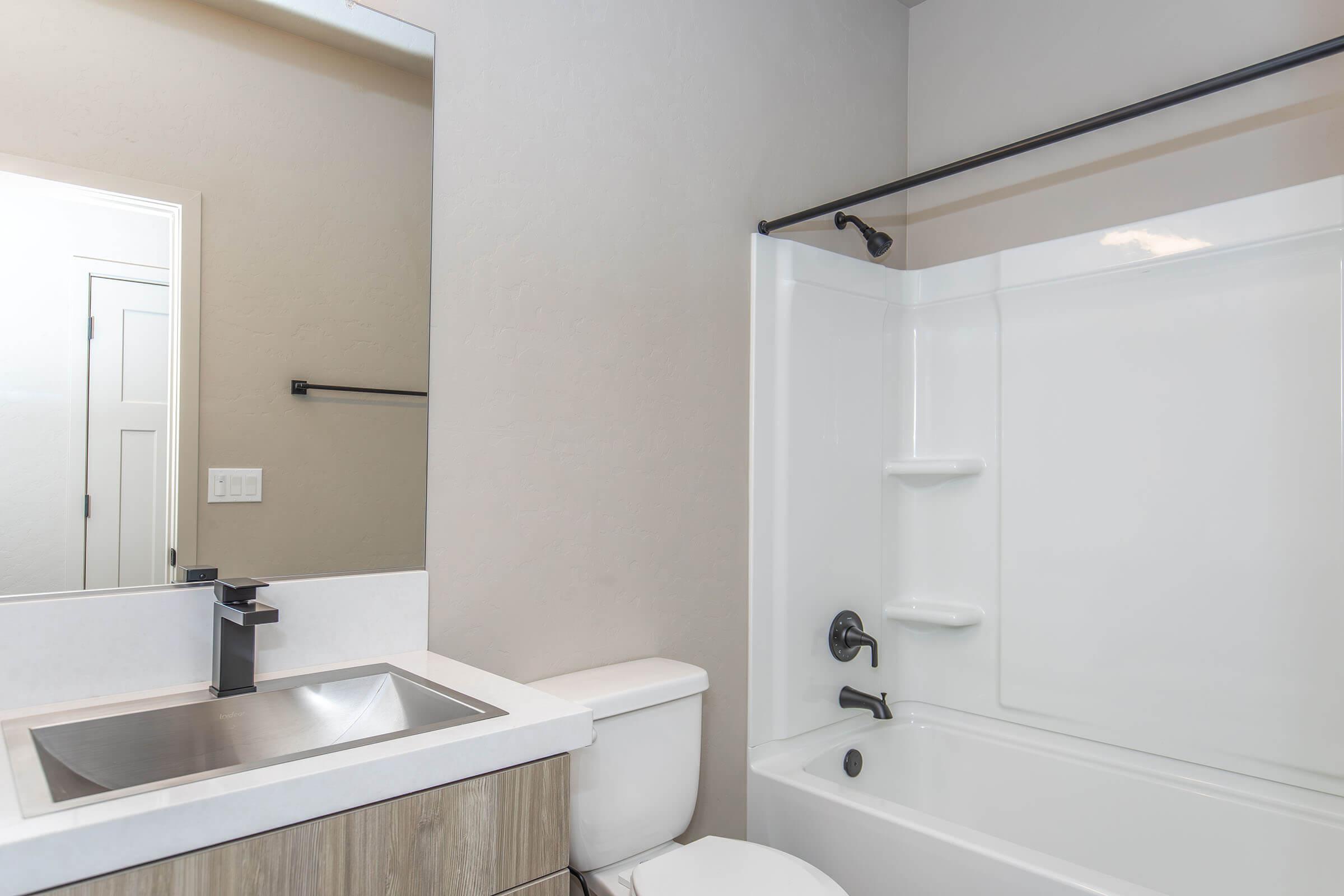 A modern bathroom featuring a white bathtub with a shower, a sleek black faucet, and a minimalistic sink area. The walls are painted in a light shade, complemented by a large mirror and simple bathroom fixtures. The overall design is clean and contemporary.