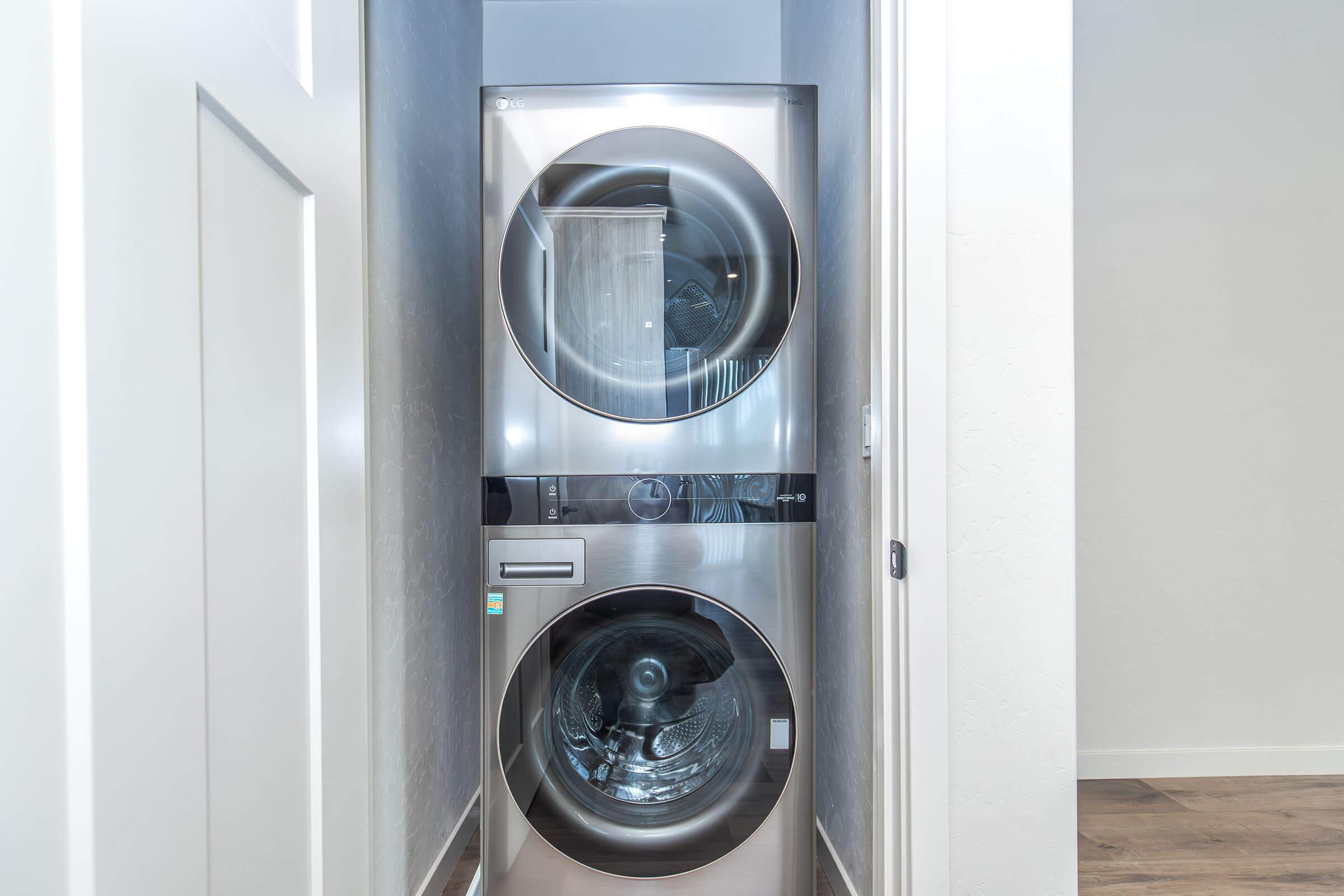 A modern stacked washer and dryer unit in a narrow laundry space, featuring sleek stainless steel finishes and transparent doors that allow visibility of the interior. The walls are painted in a light color, creating a bright and airy atmosphere.