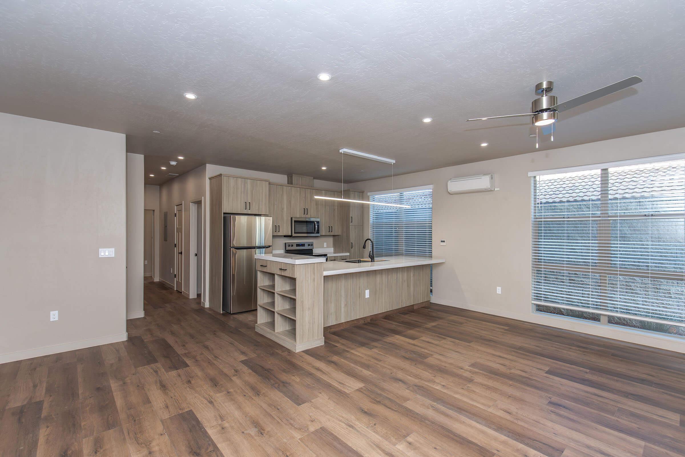 A modern, open-concept living space featuring light wood flooring, recessed lighting, and large windows. The kitchen area includes modern appliances and a central island, with a ceiling fan above. The layout seamlessly connects the living and dining areas, creating a bright and airy atmosphere.