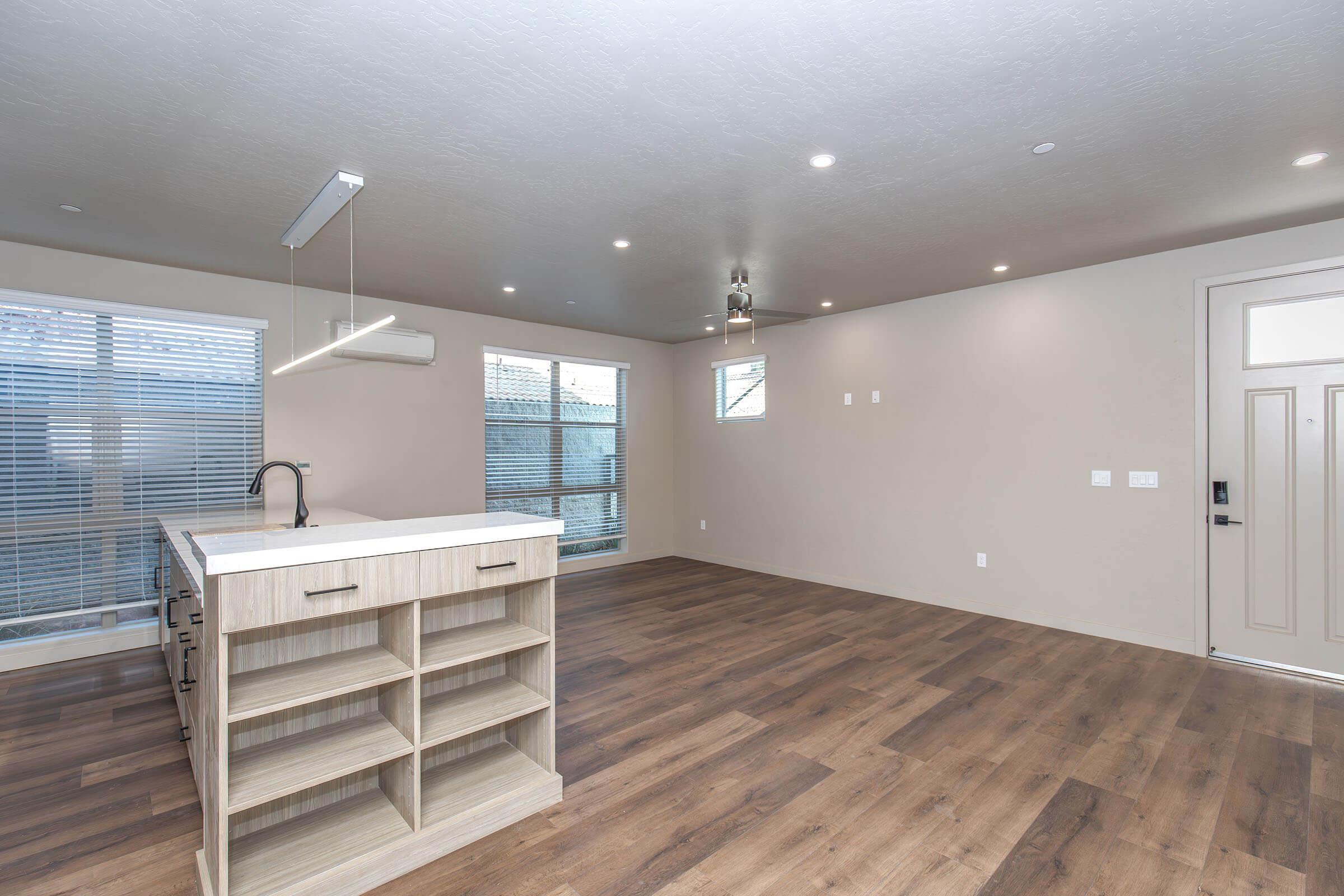 A modern, open-concept living area featuring a sleek kitchen island with shelves, large windows letting in natural light, and a spacious floor plan with light wood flooring. The walls are painted in a neutral color, creating a bright and airy atmosphere. A front door is visible on the right side.