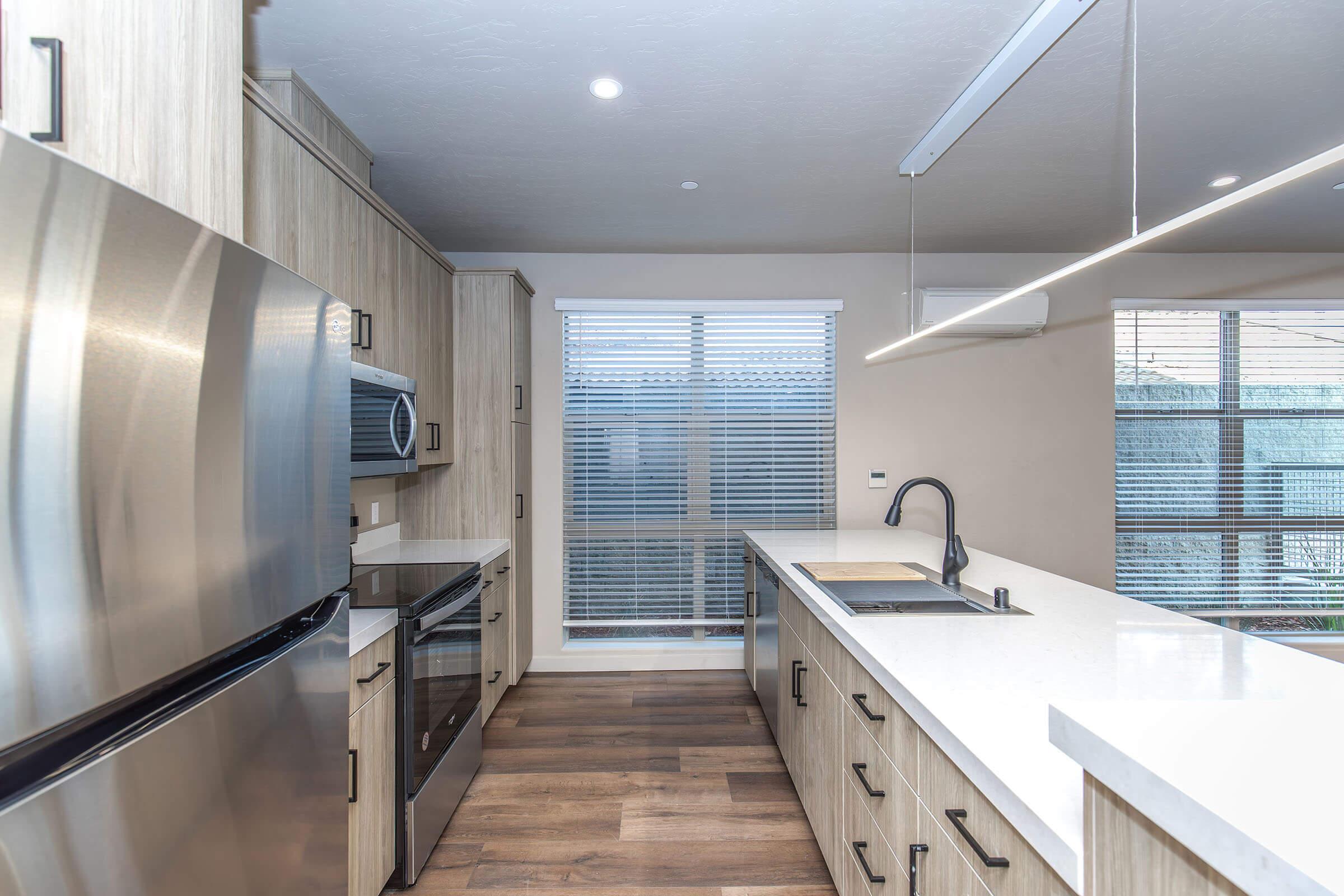 Modern kitchen featuring stainless steel appliances, including a refrigerator and oven, along with light wood cabinetry. The countertop is a sleek white surface. Natural light enters through a window with blinds, creating a bright and inviting atmosphere. The flooring is a warm wood tone.