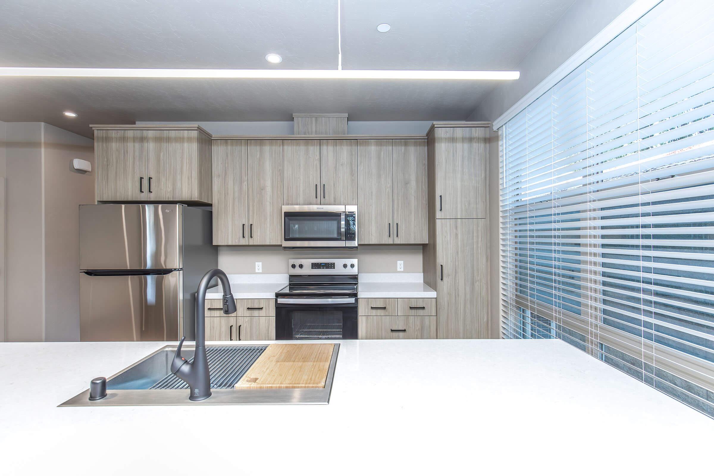 Modern kitchen featuring wooden cabinetry, stainless steel appliances including a refrigerator and oven, a sink with a sleek faucet, and a large countertop. Bright natural light streams through the blinds on the window, creating a spacious and inviting atmosphere.