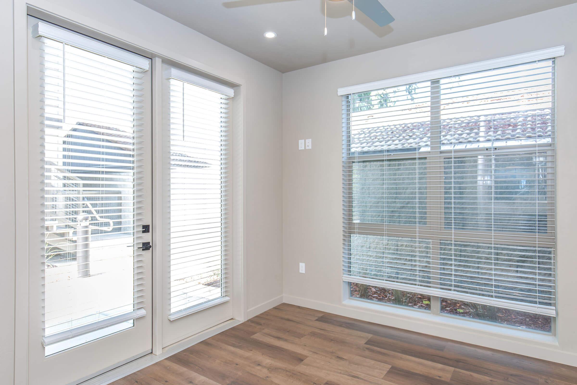 A bright interior corner of a room featuring large windows with white blinds, a door leading outside, and light-colored walls. The floor is made of wooden planks, and the overall space appears clean and modern. Natural light enters from the windows, enhancing the ambiance.
