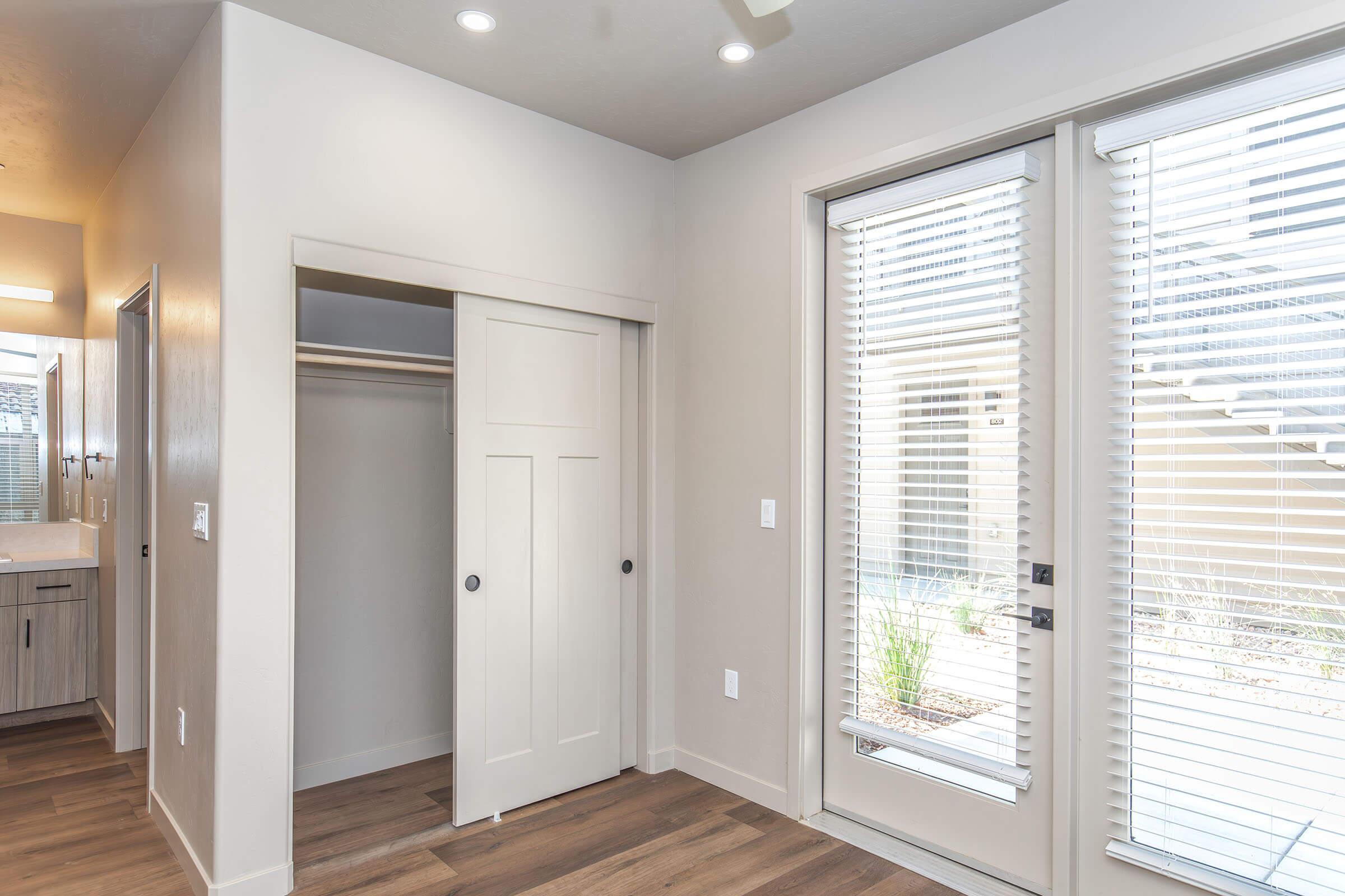 A modern interior space featuring a sliding closet door, natural wood flooring, and large glass doors leading outside. The walls are painted in a light color, and there is soft lighting from recessed ceiling fixtures. The area offers a clean and minimalist aesthetic, with a view of outdoor greenery.