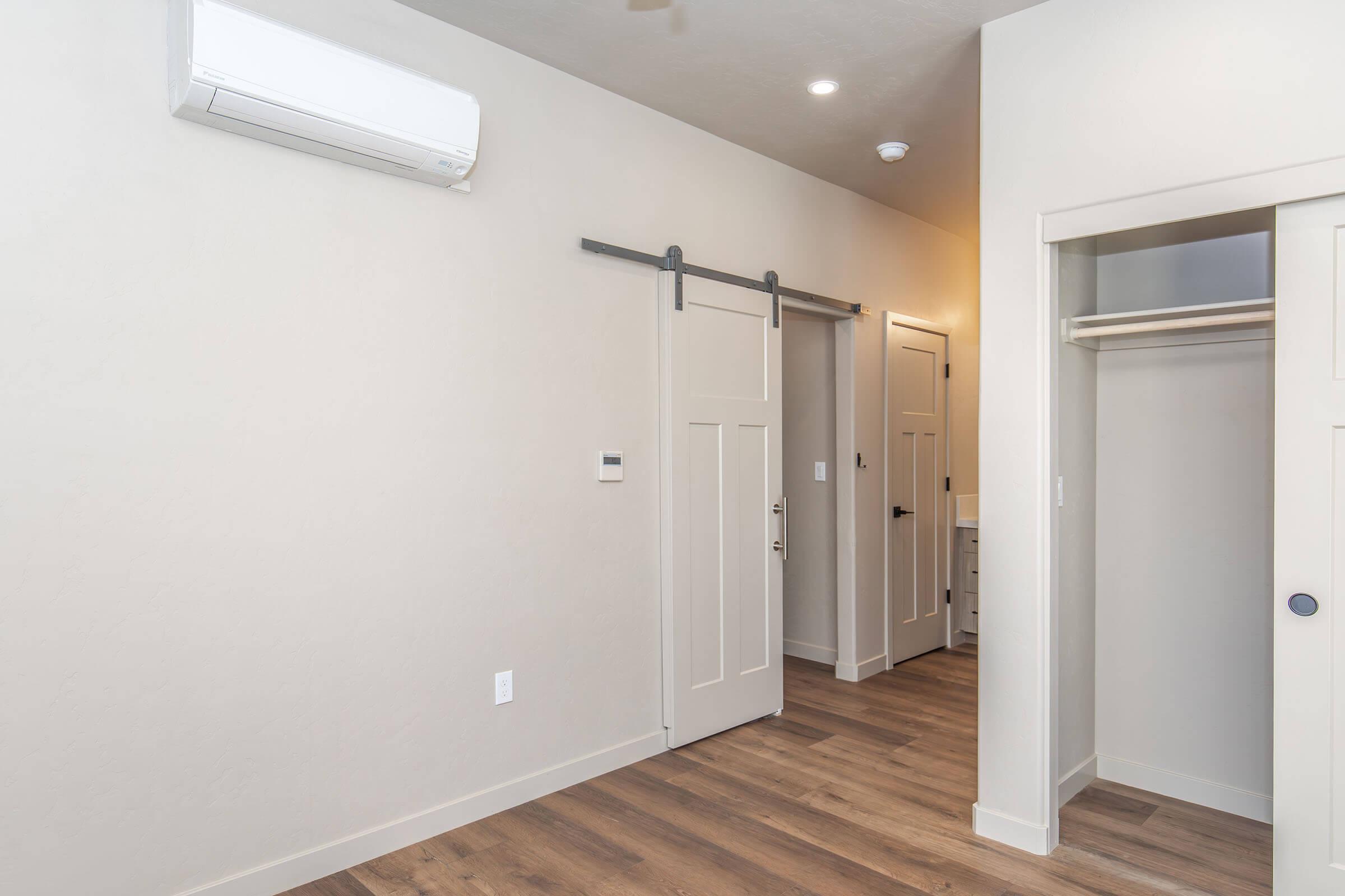 A modern interior room featuring light-colored walls, wooden flooring, and a sliding barn door. An air conditioning unit is mounted on the wall, and there is a small closet with doors on the right. The room is well-lit with natural light, creating a spacious and inviting atmosphere.