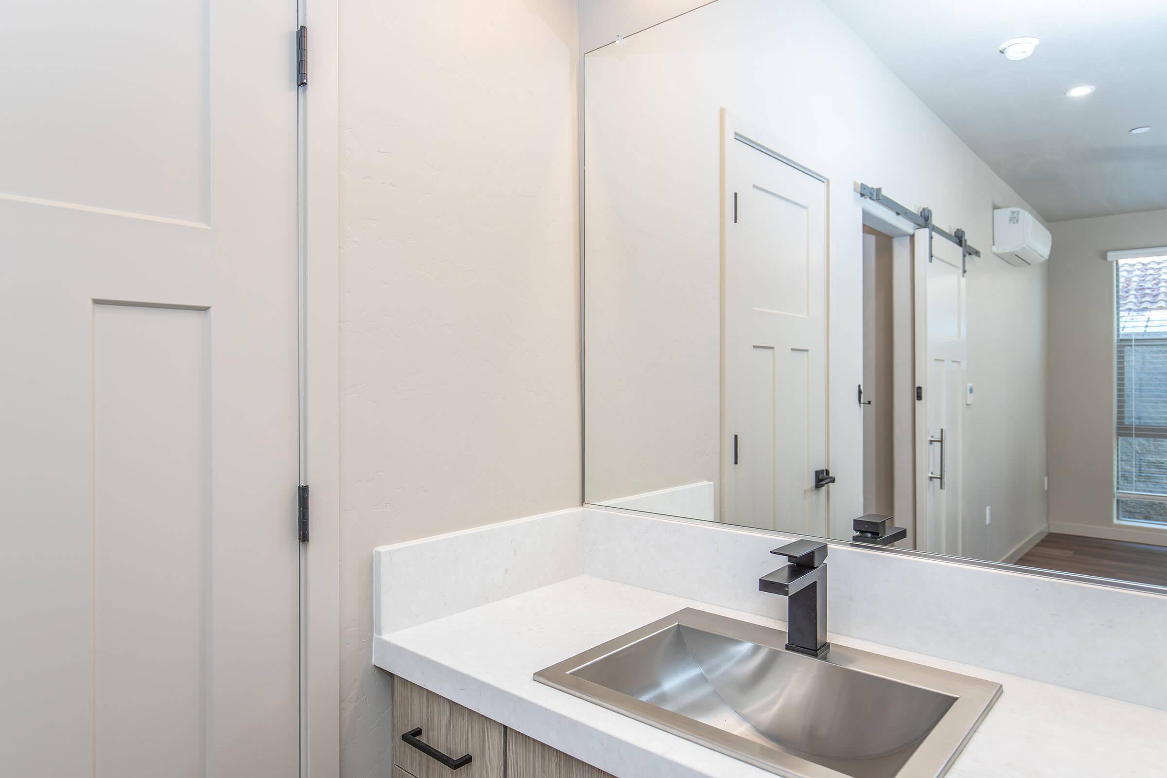A modern bathroom sink area featuring a sleek, stainless steel basin with a contemporary faucet. The walls are light-colored, and a large mirror reflects the space. There are wooden accents and a sliding door, creating a clean and minimalistic aesthetic. Natural light enters from a nearby window.