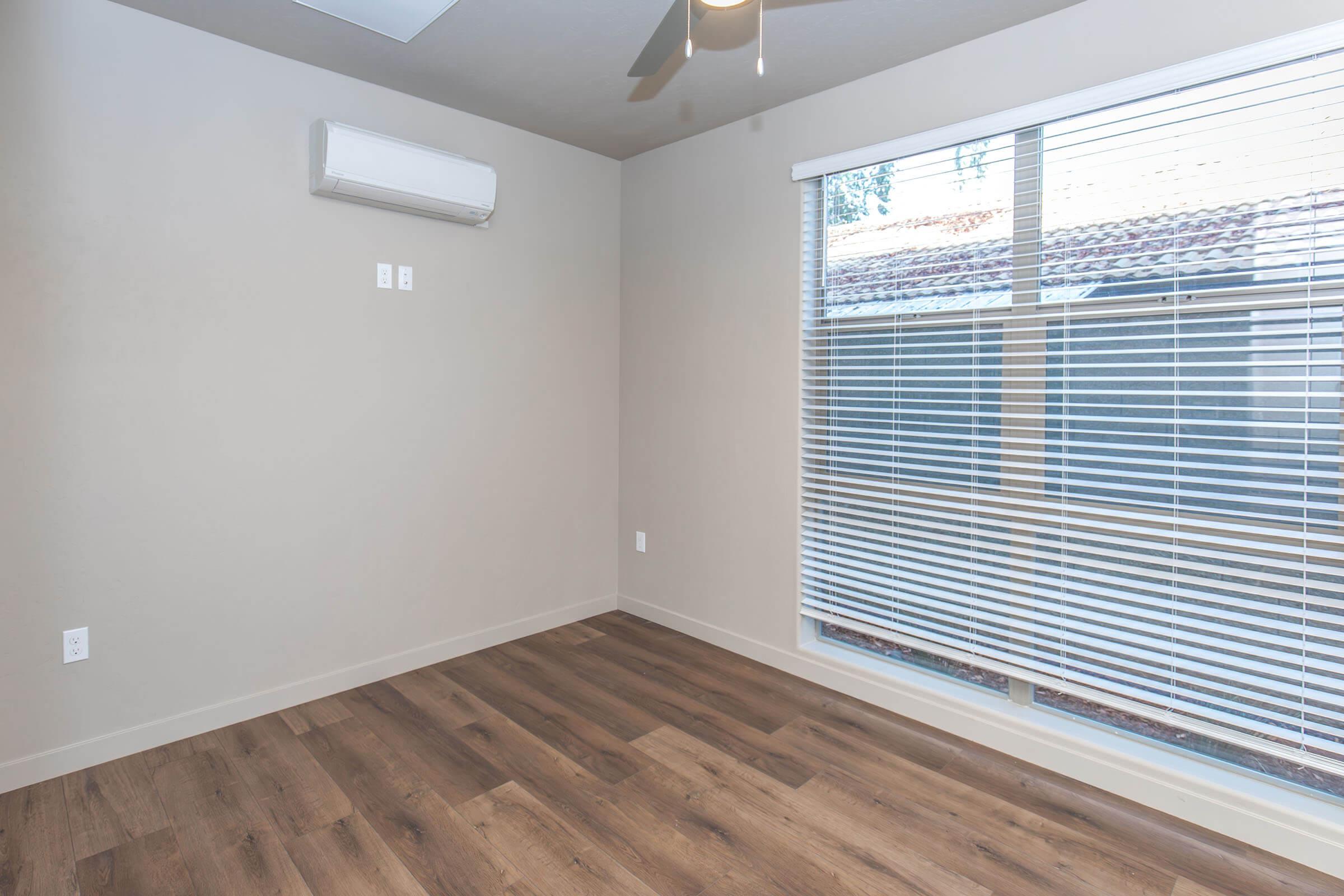 A minimalist room featuring a light gray wall, a ceiling fan, and a window with blinds. The floor is made of wooden planks, and there is an air conditioning unit mounted on the wall. The overall ambiance is bright and airy due to the natural light coming through the window.