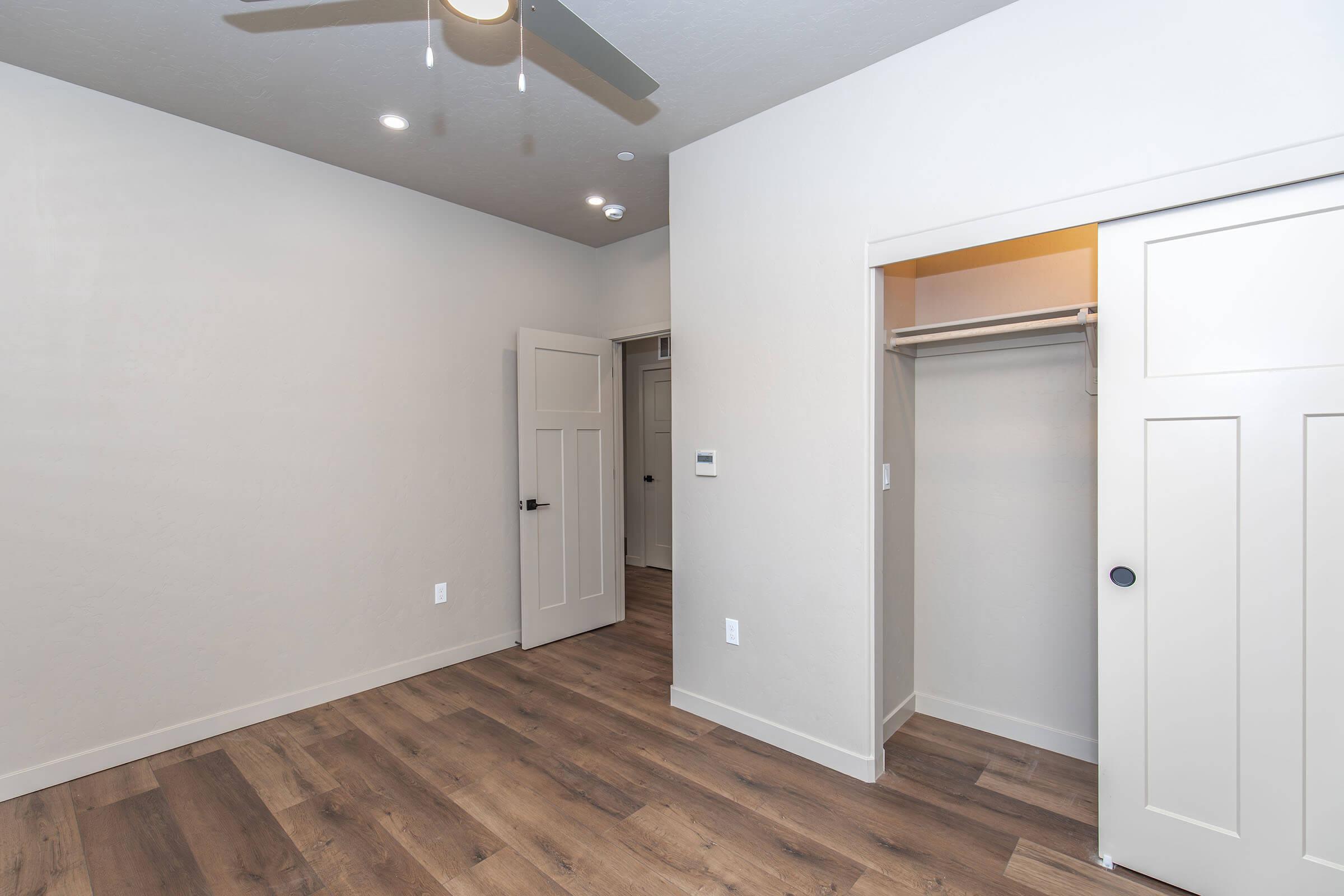 An empty room featuring light-colored walls, a ceiling fan, and wood laminate flooring. A door is visible on the left side, while a closet with sliding doors is positioned on the right. The room has a modern, minimalist design with no furniture present.