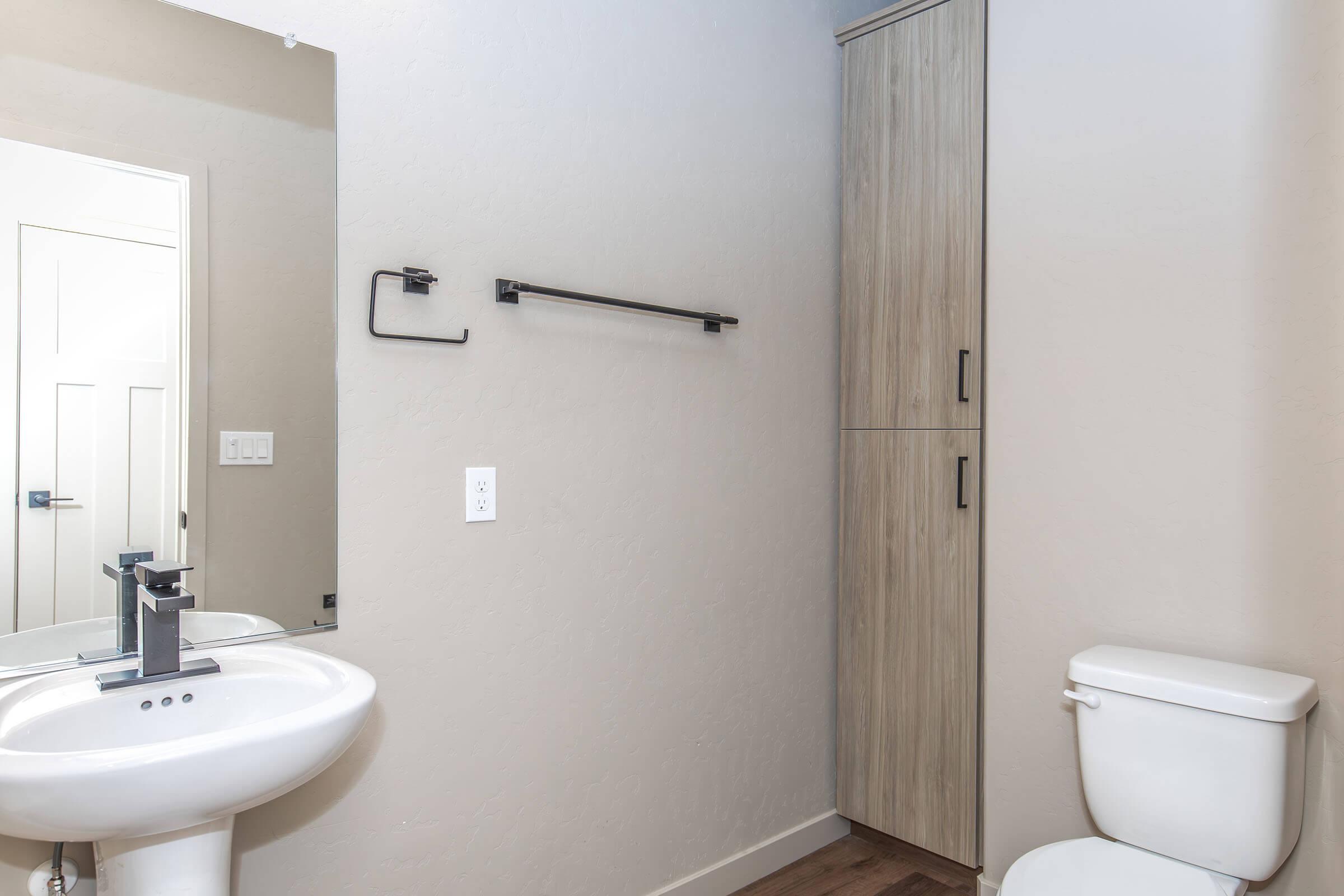 A modern bathroom featuring a white sink and faucet, a wall-mounted towel rack, a tall storage cabinet, and a white toilet. The walls are painted light colors, and the floor has wooden planks. A large mirror reflects the space, adding to the overall contemporary design.
