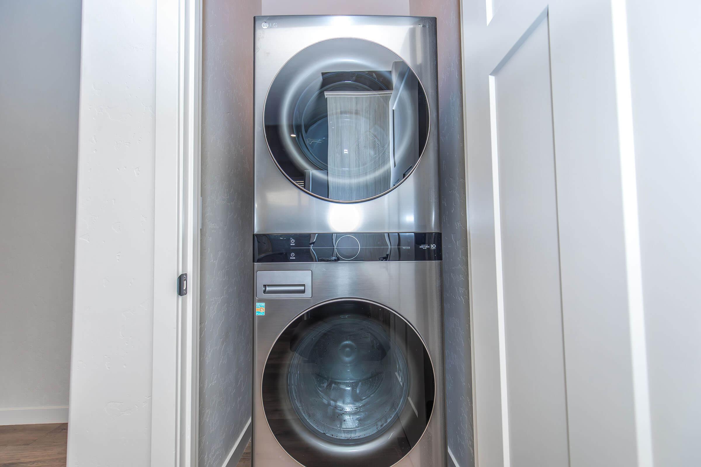A modern stacked washer and dryer in a narrow laundry space, featuring a sleek stainless steel design with front-loading doors. The setting includes light-colored walls and a door, creating a clean and organized appearance.