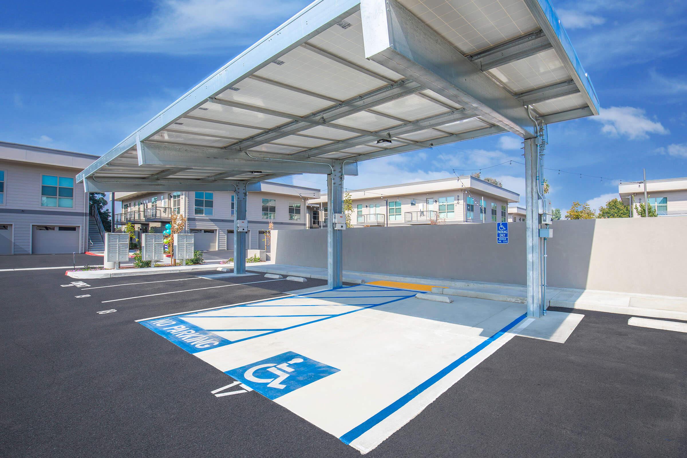 A modern electric vehicle charging station under a shaded canopy, located in a spacious parking lot. The area features designated handicapped parking spaces, clear signage, and surrounding residential buildings in the background under a blue sky.