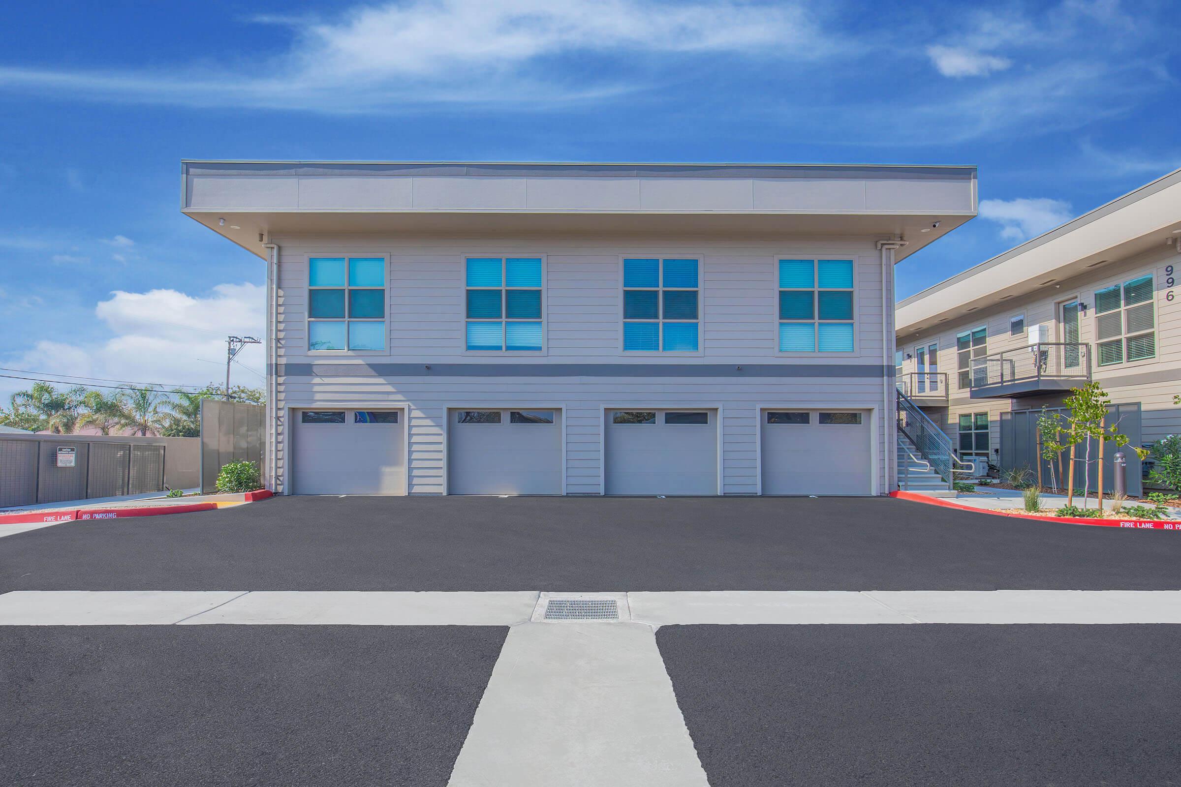 Exterior view of a modern two-story building with large windows and garage doors. The building is surrounded by a newly paved parking area and there are clear blue skies in the background. Nearby structures are visible, adding to the contemporary aesthetic.