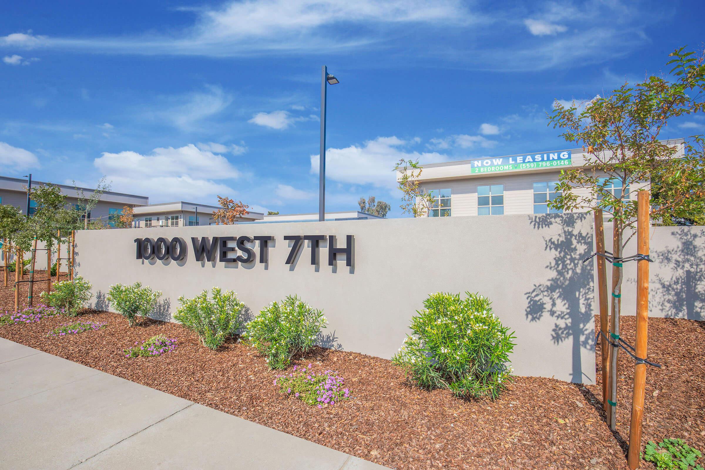 Modern exterior view of a property sign reading "1000 WEST 7TH," surrounded by landscaped plants and flowers. Clear blue skies in the background and a building with a "NOW LEASING" sign visible in the distance. The area features clean pathways and decorative mulch.