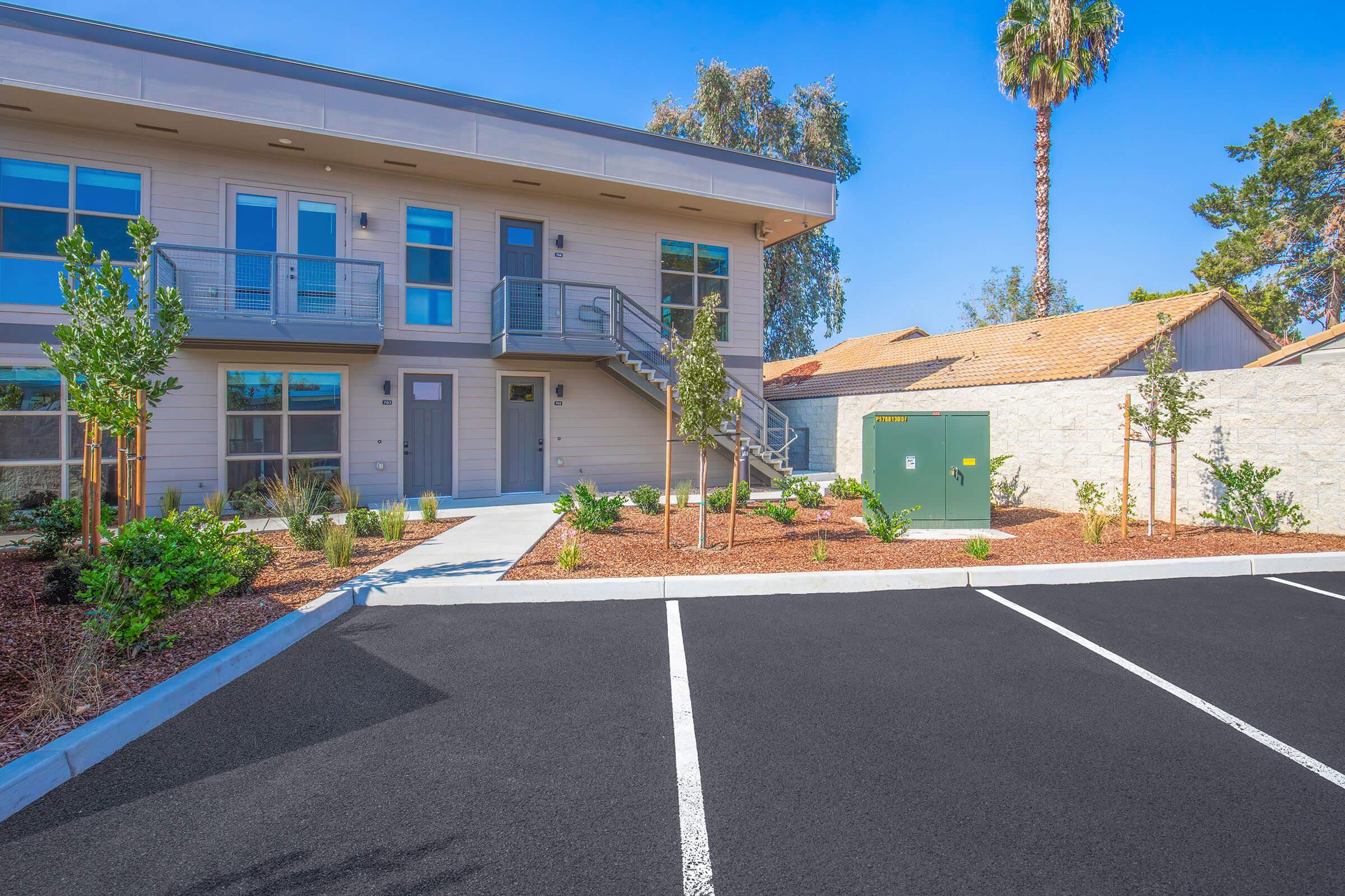 A modern two-story apartment building with a light-colored exterior, surrounded by landscaped greenery and gravel. In front is a paved parking area with several empty spaces. Palm trees and nearby structures complete the scene under a clear blue sky.