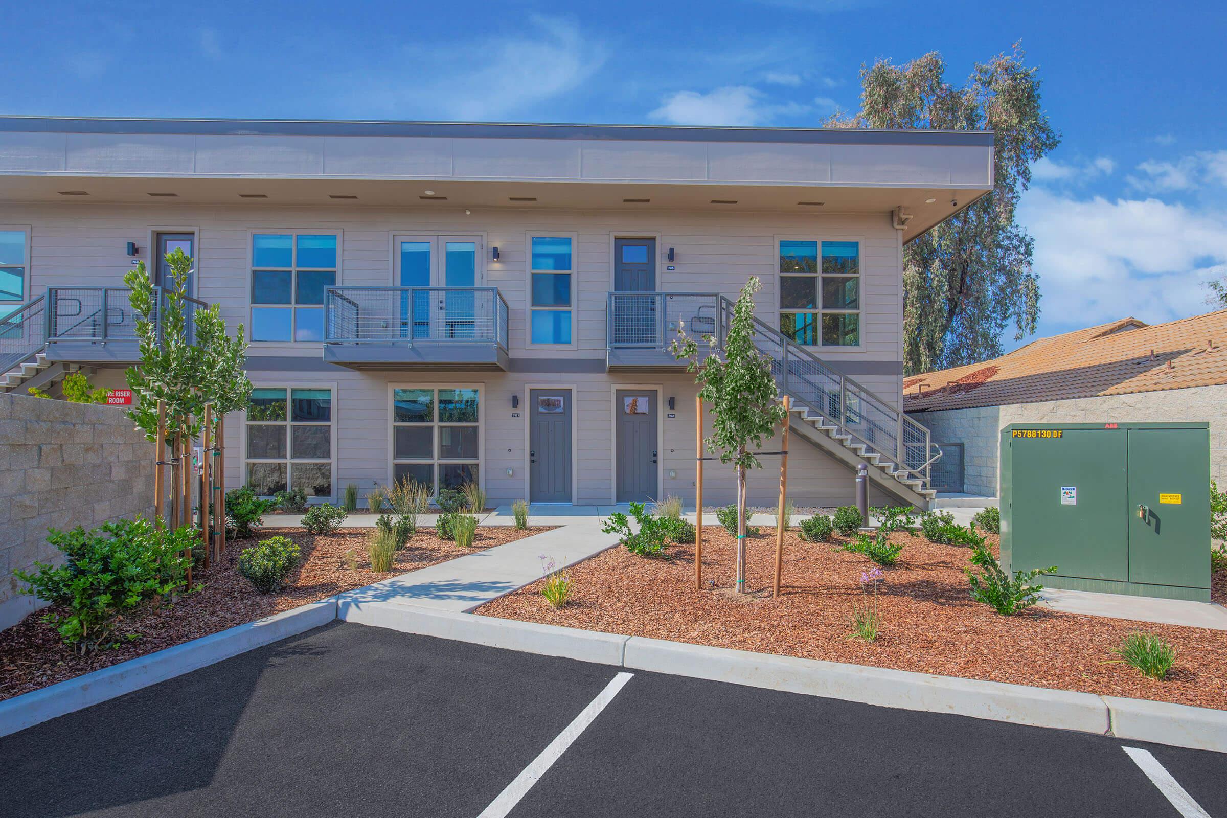 A modern two-story residential building with a minimalist design. The facade features large windows and a staircase leading to the upper floor. Well-maintained landscaping includes shrubs and small trees alongside a paved pathway. Parking spaces are visible in the foreground.