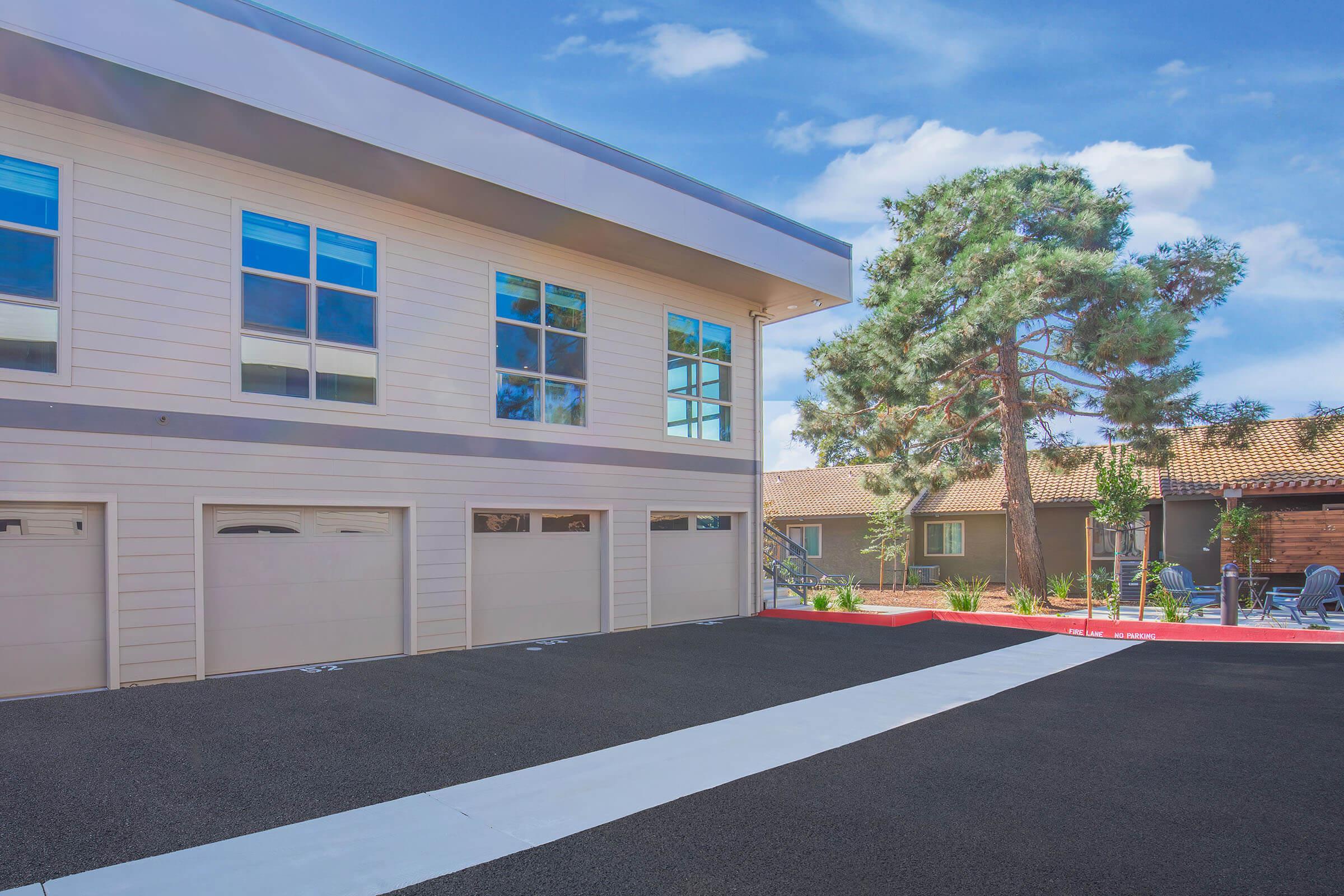 A modern building exterior featuring large windows, a gray facade, and three garage doors. The surrounding area includes landscaped greenery, a mature tree, and a paved driveway. In the background, additional buildings and outdoor seating are visible, set against a clear blue sky.