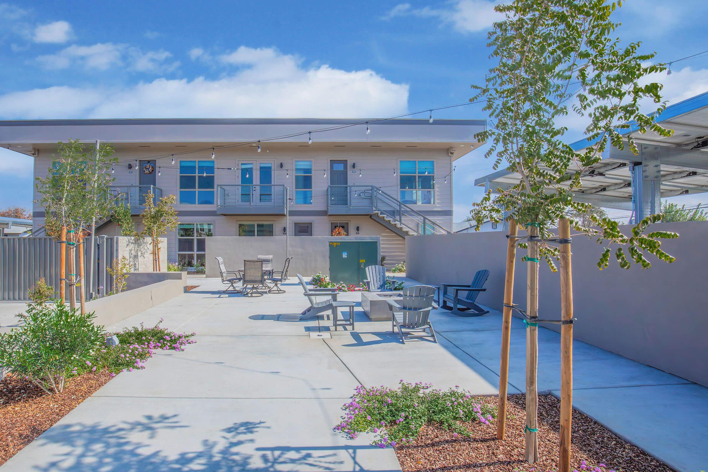 A modern outdoor space featuring a two-story building with a staircase. Stylish seating areas with gray chairs and tables are surrounded by landscaped gardens with flowering plants and young trees. The area is bright and inviting, under a clear blue sky.