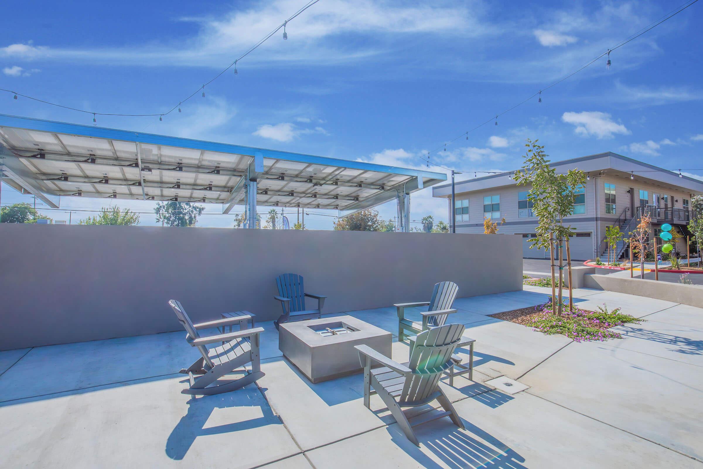 Outdoor seating area featuring several modern chairs surrounding a fire pit, with a large solar panel structure overhead. In the background, a contemporary building and landscaped greenery are visible under a clear blue sky with scattered clouds.