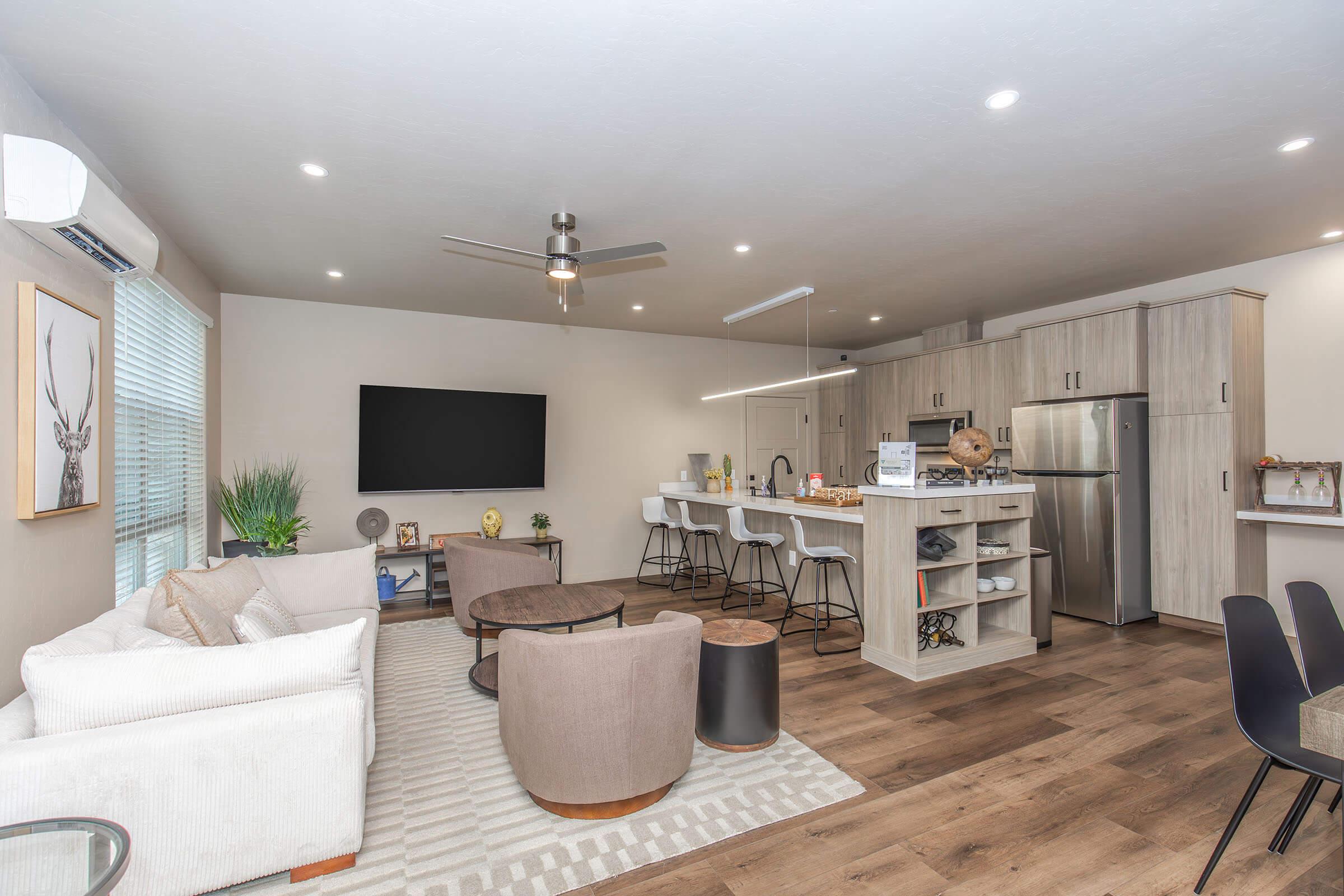 Modern living space featuring a cozy seating area with a white sofa and round coffee table. A kitchen area with a stainless steel fridge and bar stools sits in the background. There’s a large flat-screen TV on the wall, soft lighting, and a decorative plant, creating a warm, inviting atmosphere.