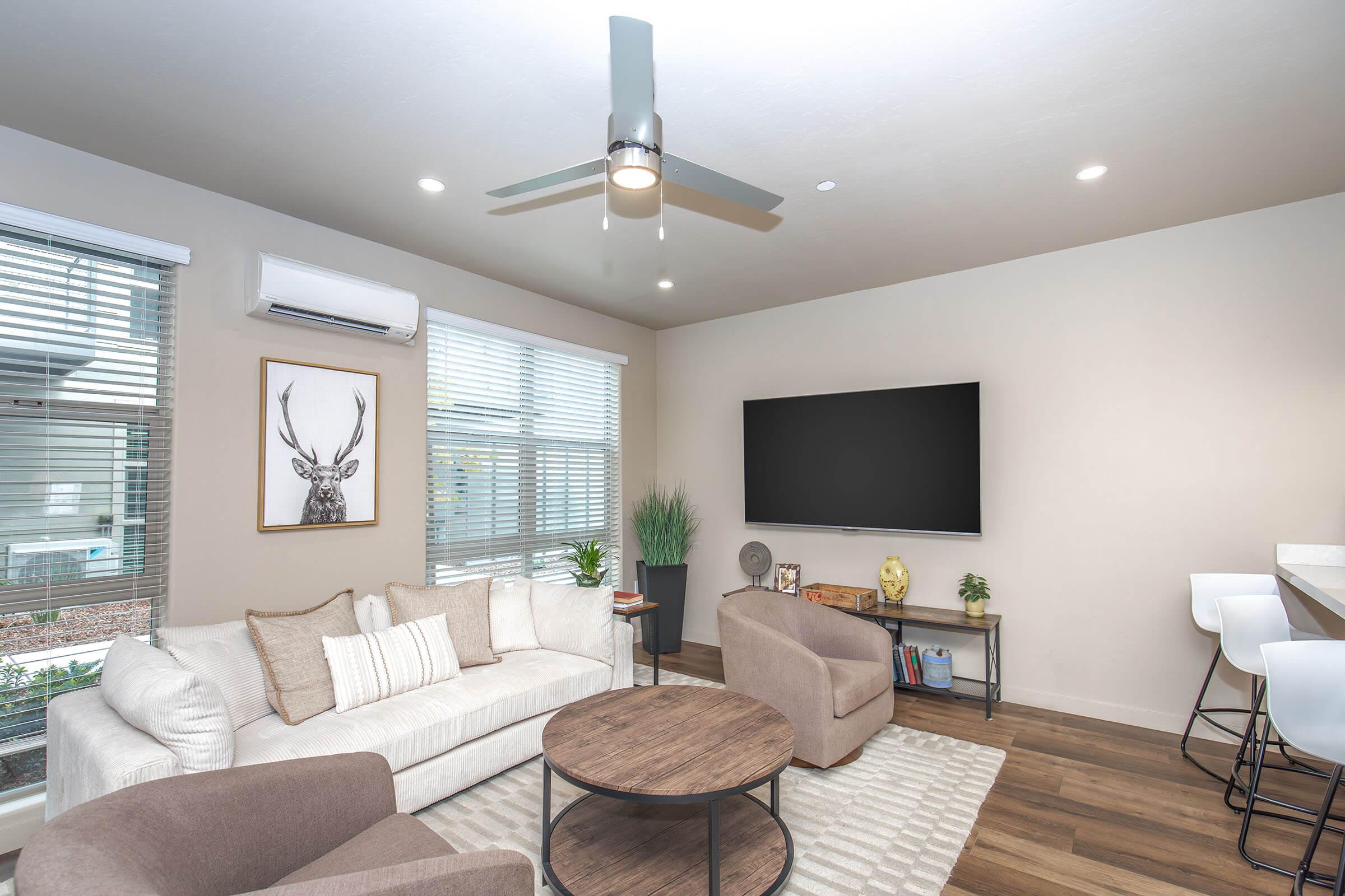 A modern living room featuring a light-colored sofa with decorative cushions, two brown armchairs, a round coffee table, and a large flat-screen TV mounted on the wall. There are potted plants, a framed deer artwork, and large windows allowing natural light to illuminate the space.