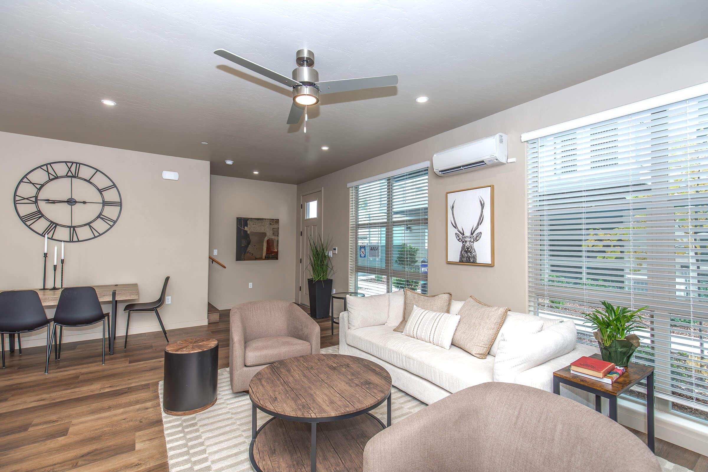 Modern living room featuring a neutral color palette, a comfortable sofa with decorative pillows, a round coffee table, and stylish armchairs. A large wall clock and framed artwork add character to the space, while large windows allow natural light to fill the room, complemented by indoor plants and minimalistic decor.