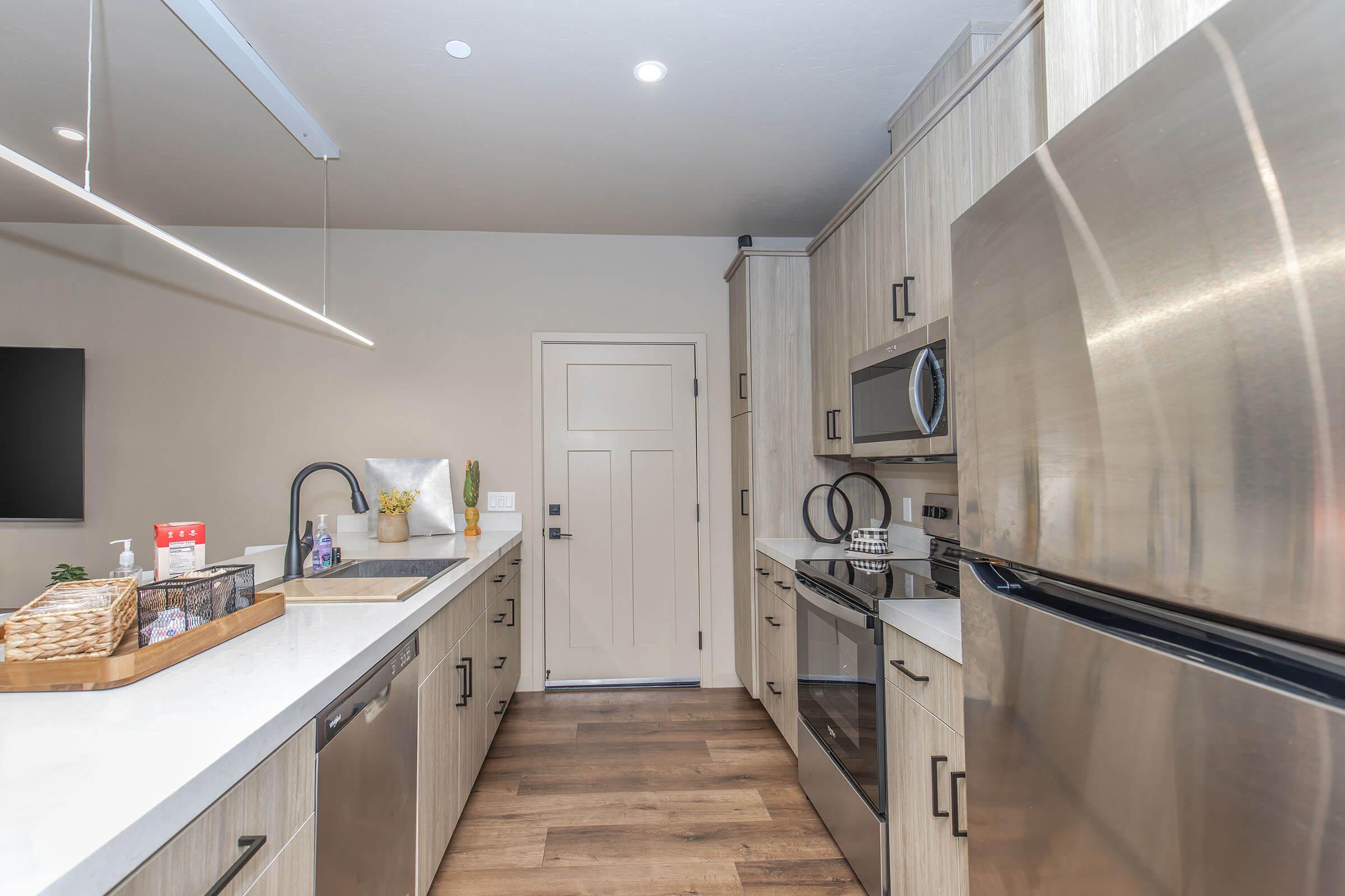 A modern kitchen featuring light wooden cabinetry, stainless steel appliances, and a white countertop. A black faucet is positioned over the sink, and a small tray with kitchen essentials is on the counter. A door leads to another area, and there is a wall-mounted TV nearby.