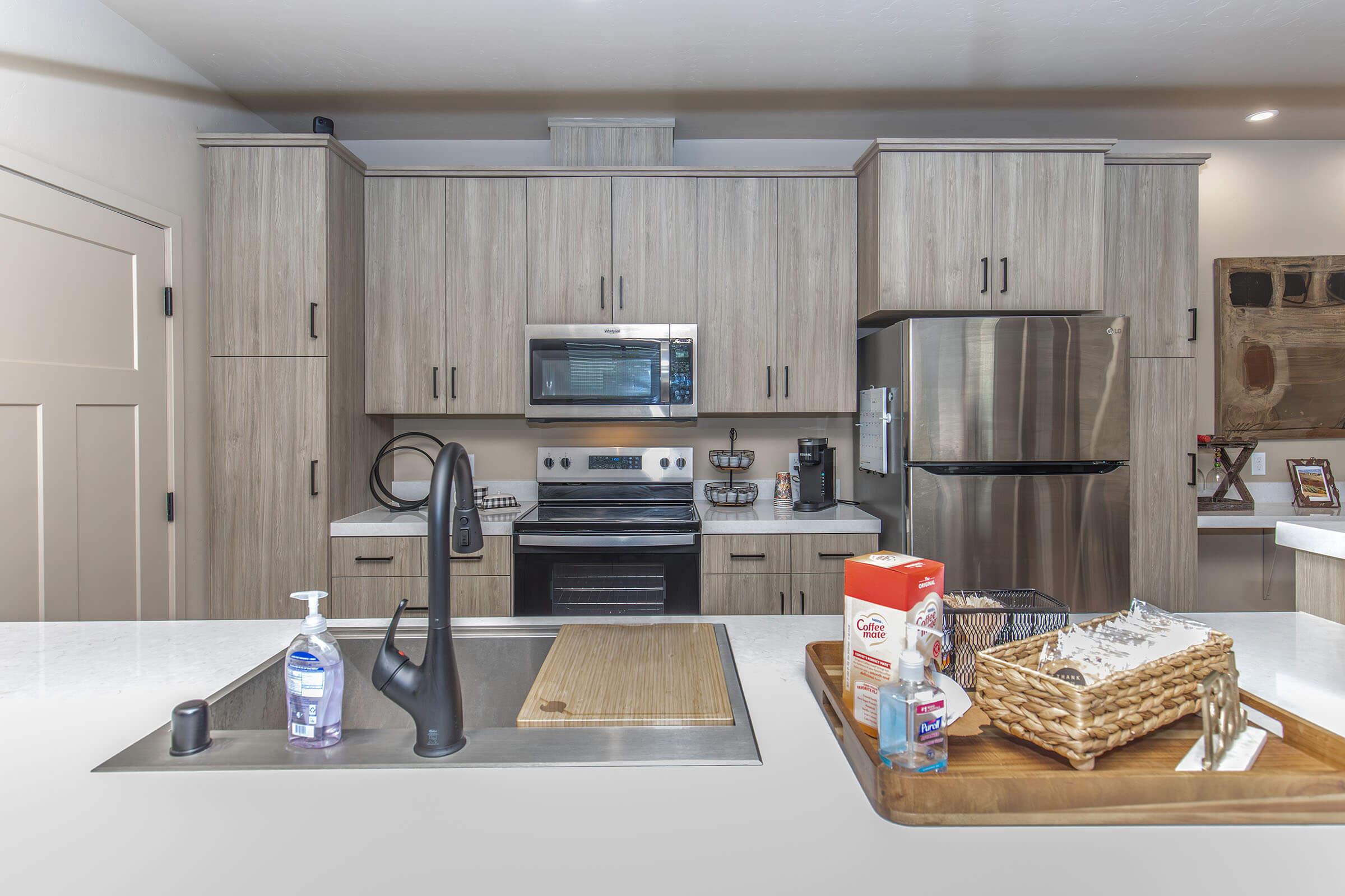 Modern kitchen with wooden cabinetry, a stainless steel refrigerator, and an oven. A sink with a black faucet is in the foreground, next to a cutting board and a decorative basket. The countertop features a soap dispenser and various kitchen items, creating a tidy and functional space.