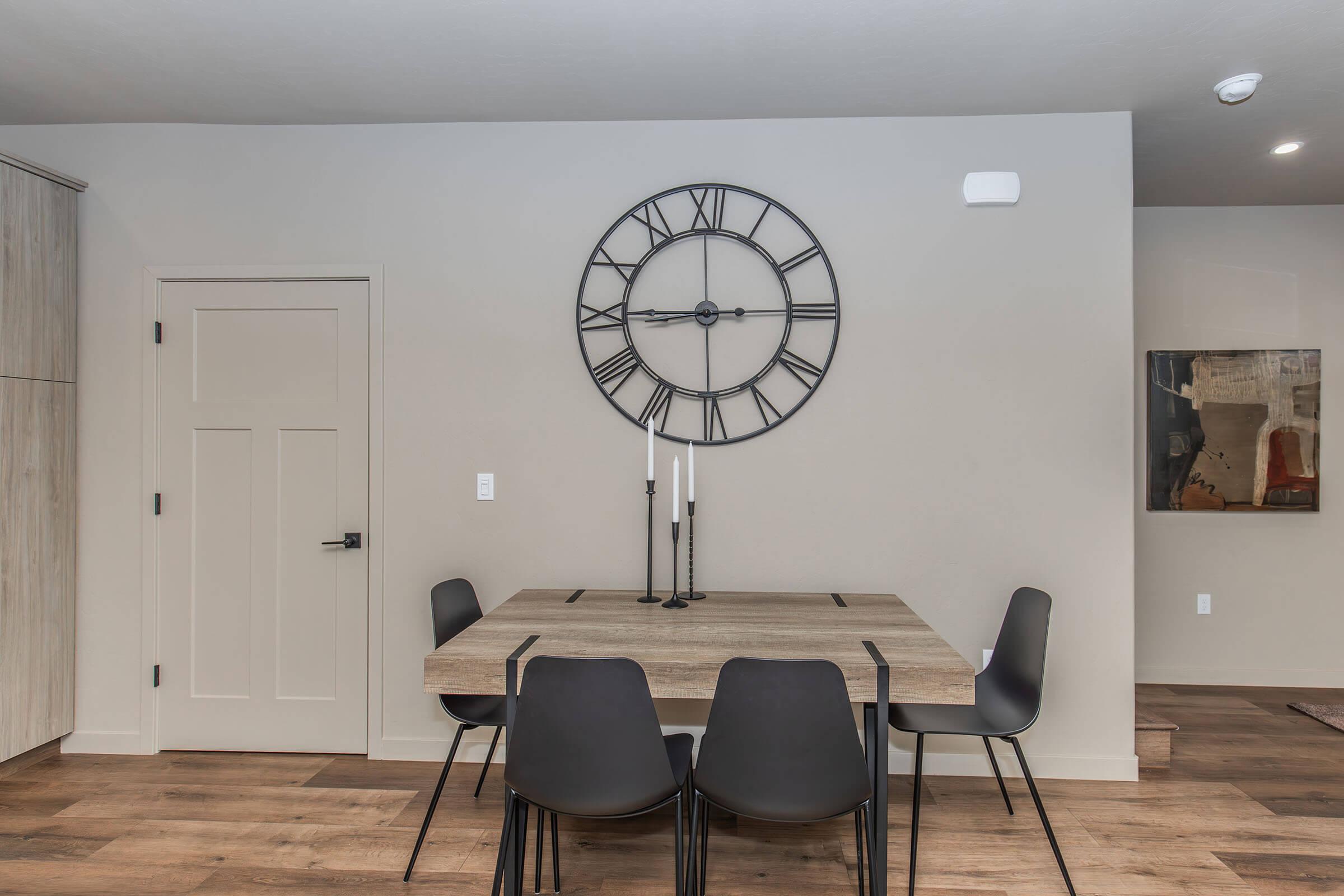 A modern dining area featuring a wooden table with four black chairs. A large wall clock in black metal hangs above the table. The walls are painted a light neutral color, complemented by a piece of abstract artwork to the right. Natural light floods the space, enhancing its contemporary feel.