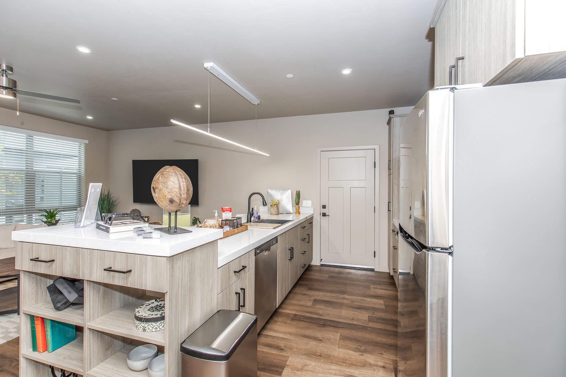 Modern kitchen with light wood cabinetry, a large island featuring a white countertop, and stainless steel appliances. There is a circular decorative globe on the counter, a dining area with a view of a window, and a door leading outside. The space is well-lit and stylishly designed.