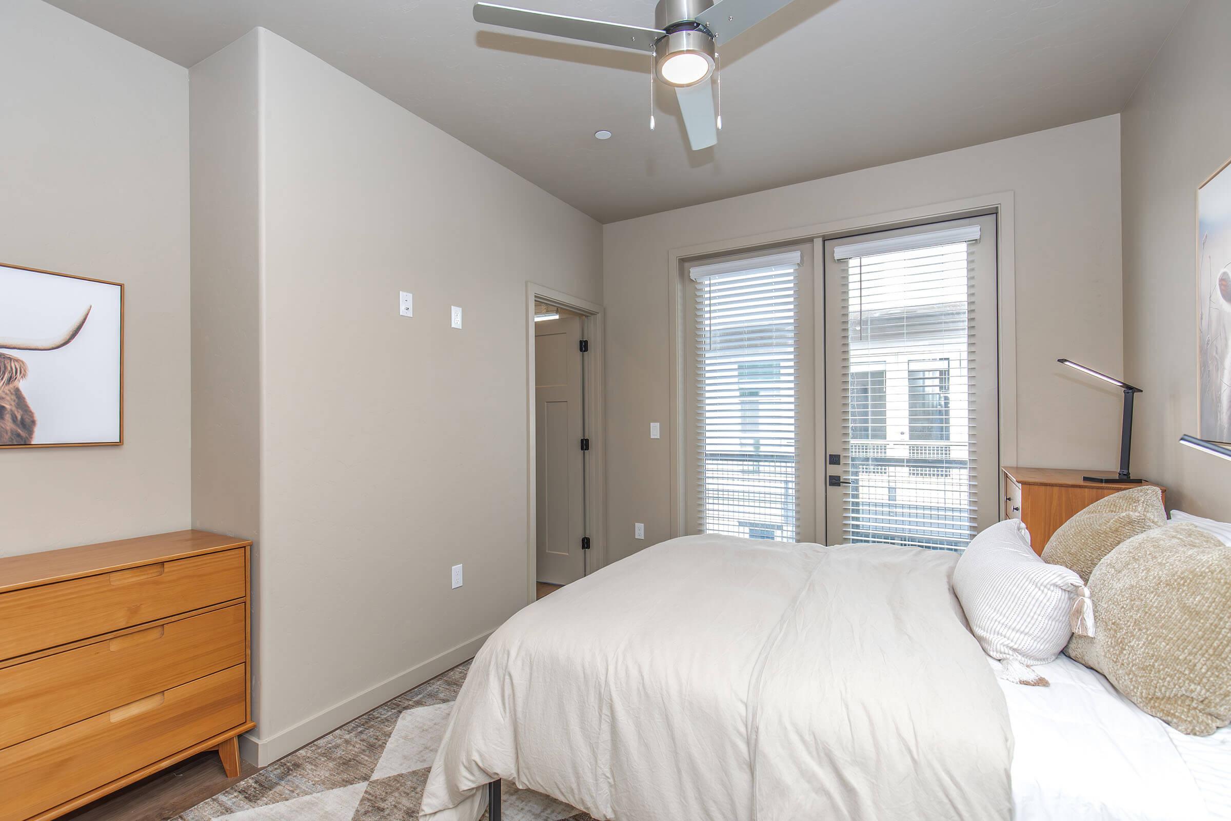 Bright and modern bedroom featuring a neatly made bed with beige and white linens. A wooden dresser stands against the wall, and large windows allow natural light to fill the room. The walls are painted in light tones, creating a serene atmosphere. A ceiling fan is also visible.