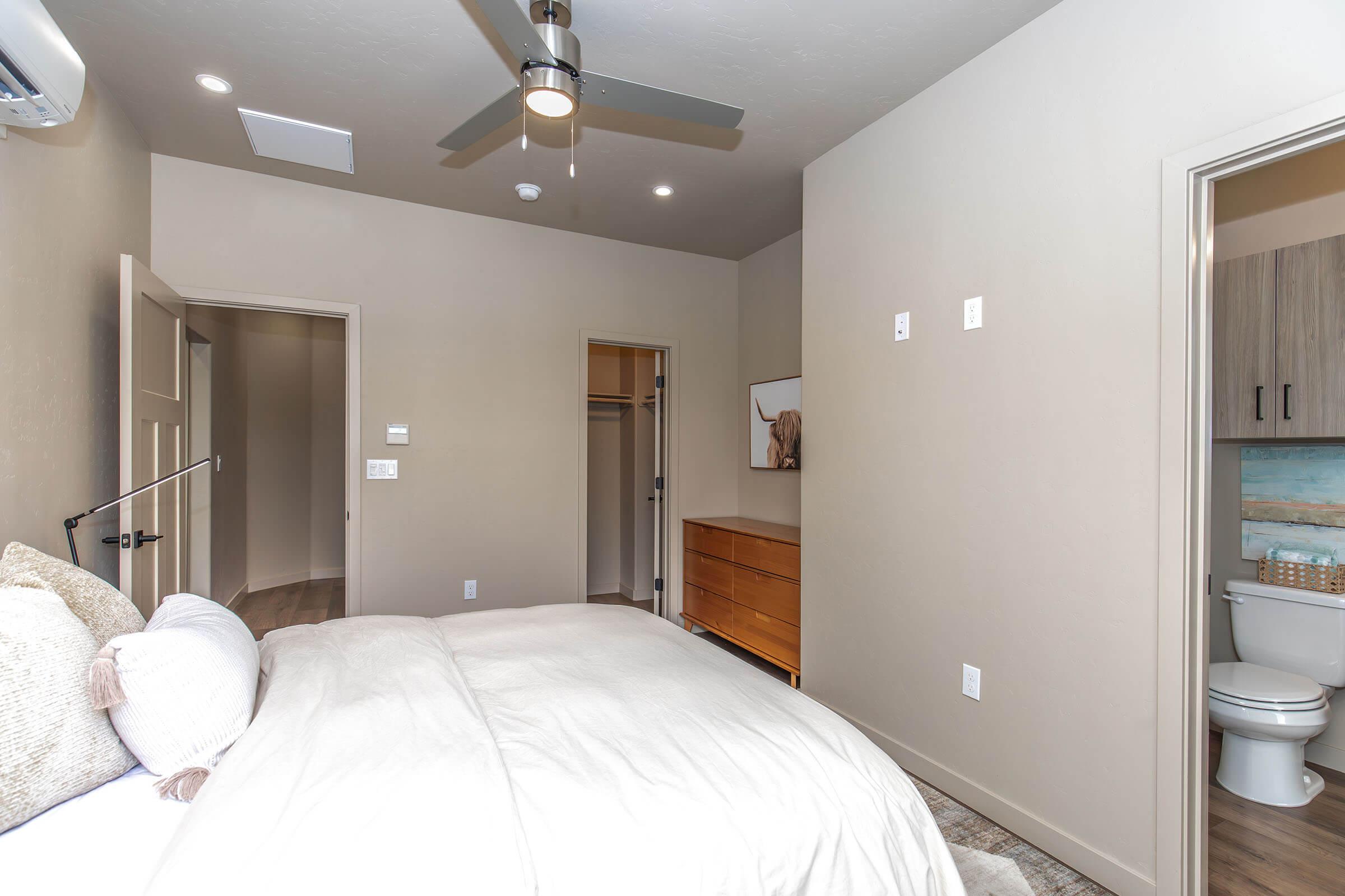 A modern bedroom featuring a neatly made bed, a dresser, and a ceiling fan. A door leading to a bathroom is visible on the right, while a closet space can be seen to the left. The walls are painted in a neutral color, contributing to a calm atmosphere. Natural light enters the room, enhancing the spacious feel.