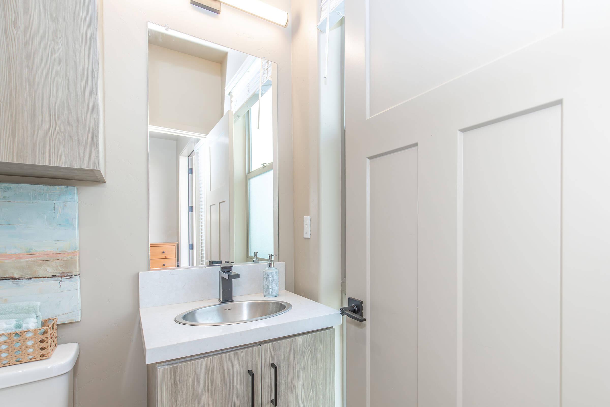 A well-lit bathroom featuring a small sink with a mirror above it. The sink area includes minimalist decor and a container on the countertop. A doorway leads to another room, with light-colored walls and a wooden cabinet visible in the background.