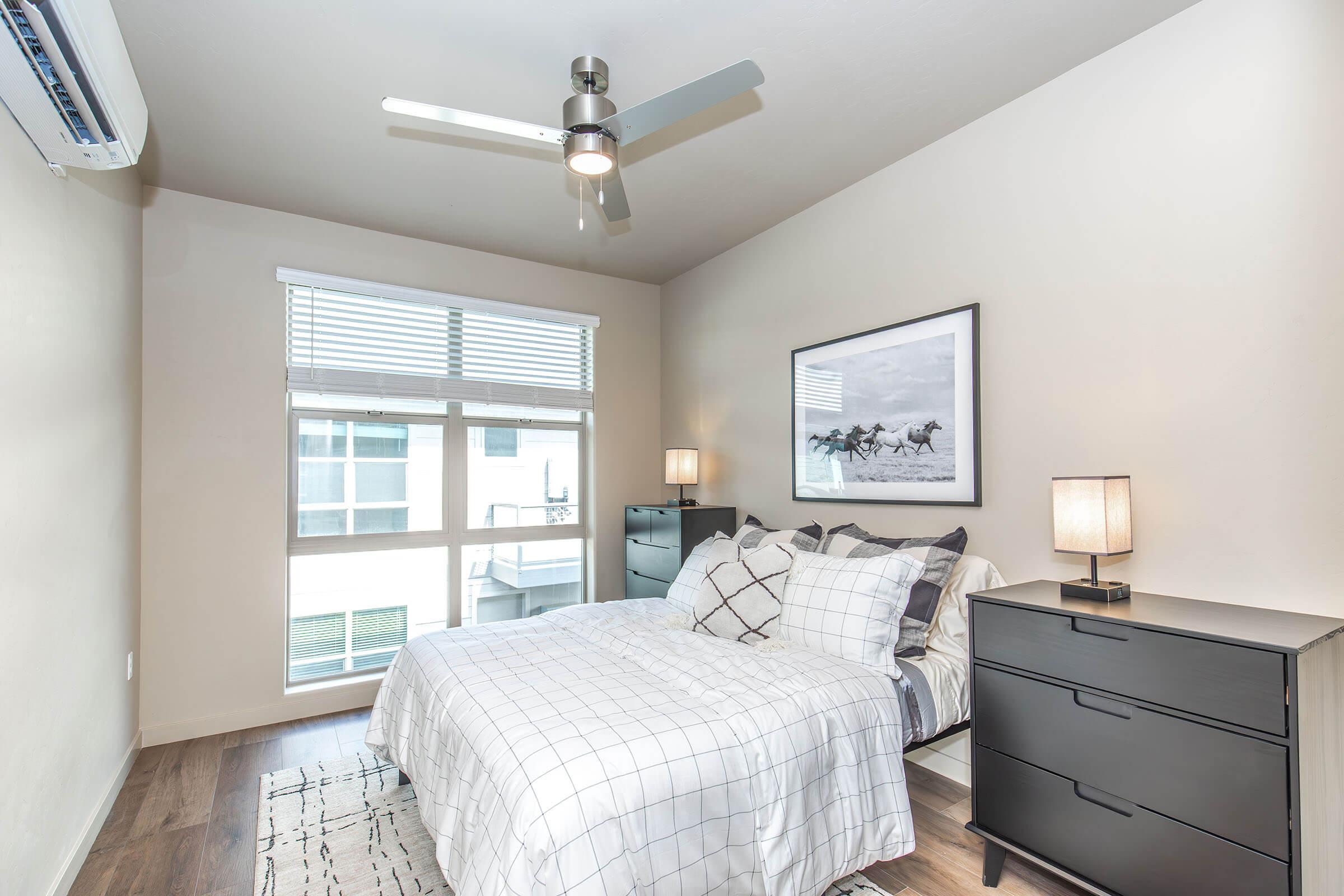 Cozy bedroom featuring a bed with a checkered duvet, black dresser, and two bedside lamps. There's a large window with white blinds allowing natural light, and a framed black-and-white horse photograph on the wall. The room has wooden flooring and a simple, modern design.