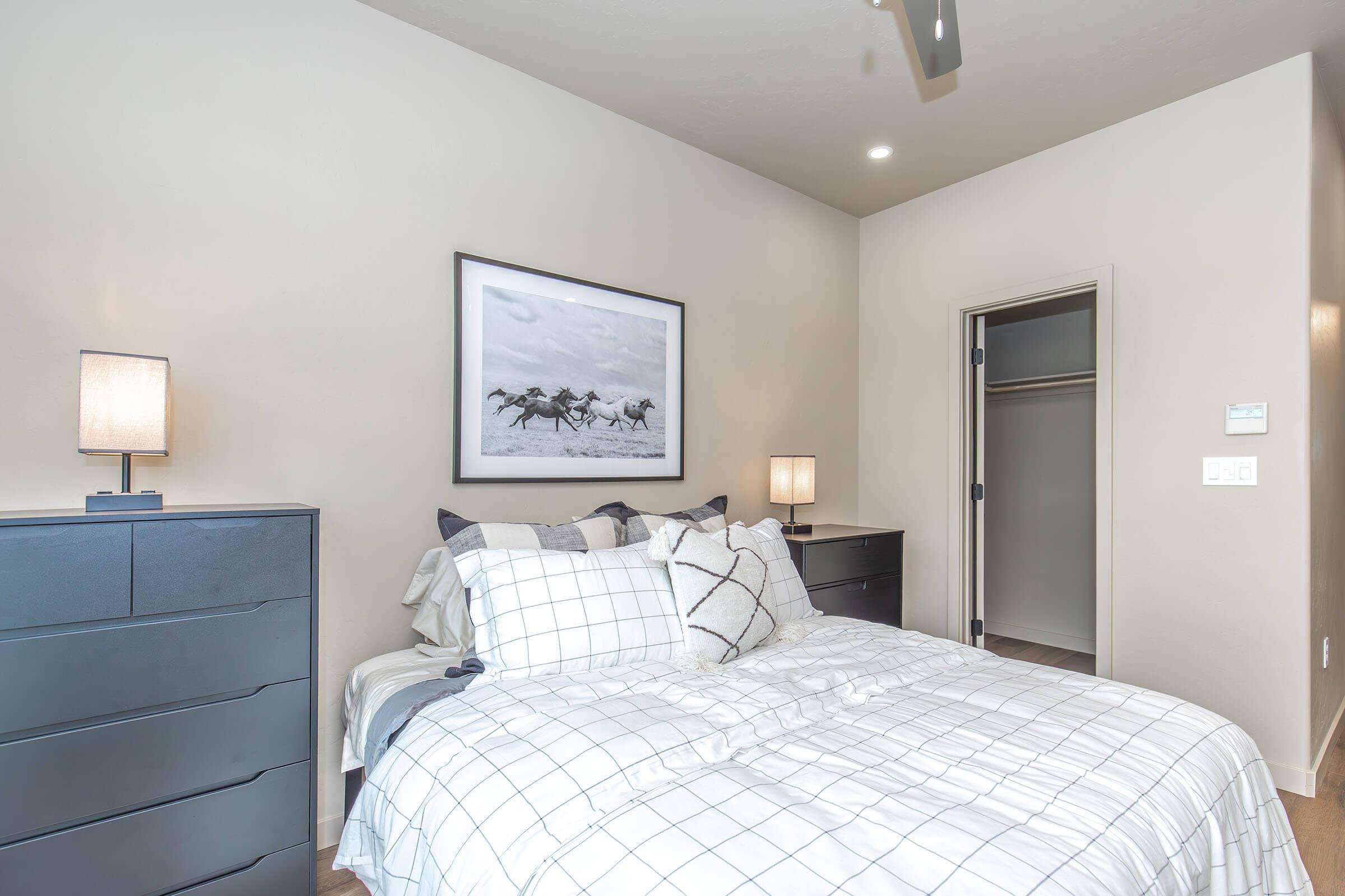 A cozy bedroom featuring a neatly made bed with a checkered white and gray comforter and decorative pillows. There are two bedside lamps on black nightstands, and a large framed black-and-white photograph of horses on the wall. A closet door is visible in the background. The walls are a soft cream color.