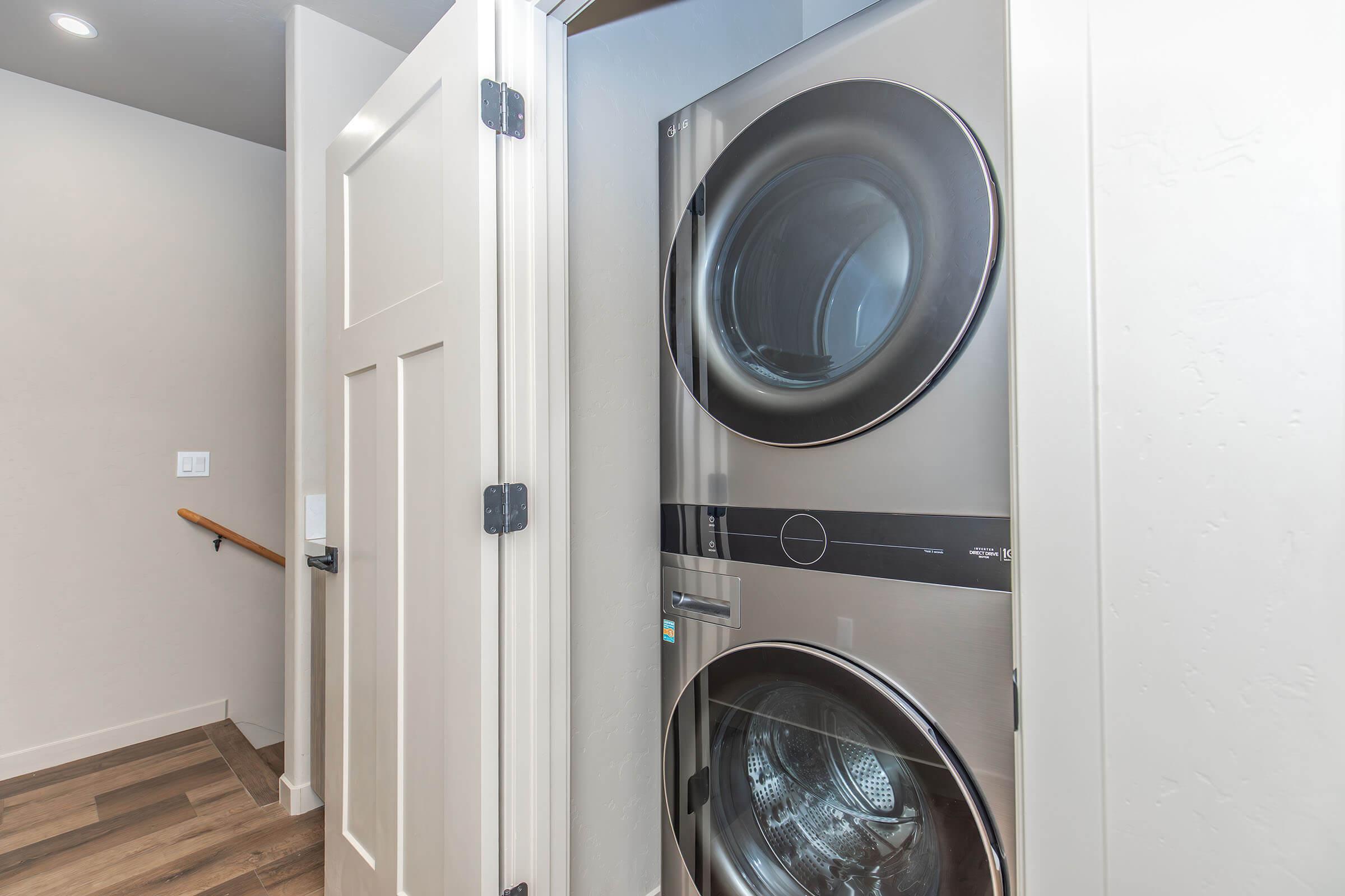 Stacked washer and dryer unit in a laundry room, partially visible through an open door. The walls are painted in a light color, and there is a wooden staircase nearby. The flooring is made of polished wood, contributing to a modern interior design aesthetic.